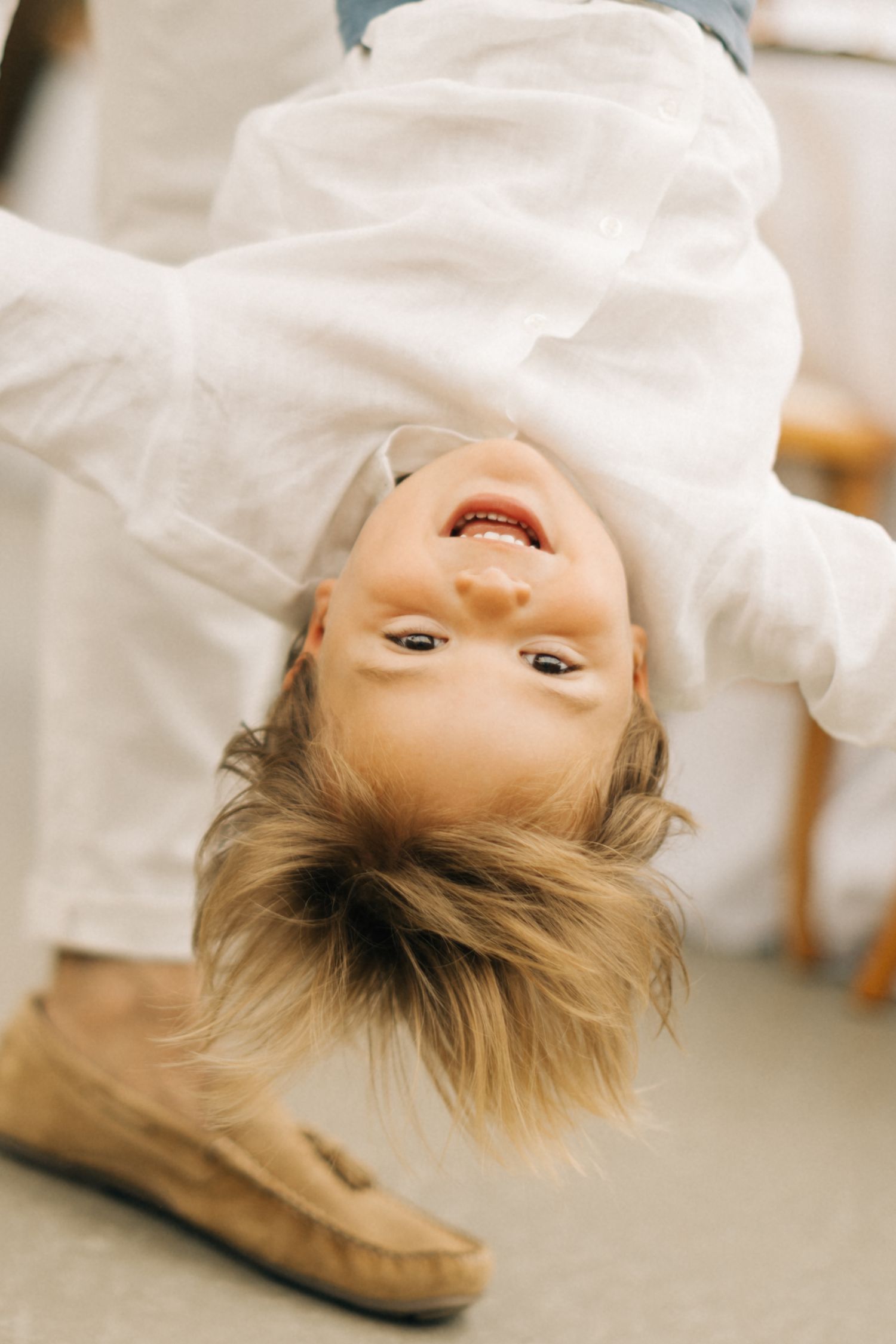 Child playfully hanging upside down in a white shirt and boots on a carpeted floor.