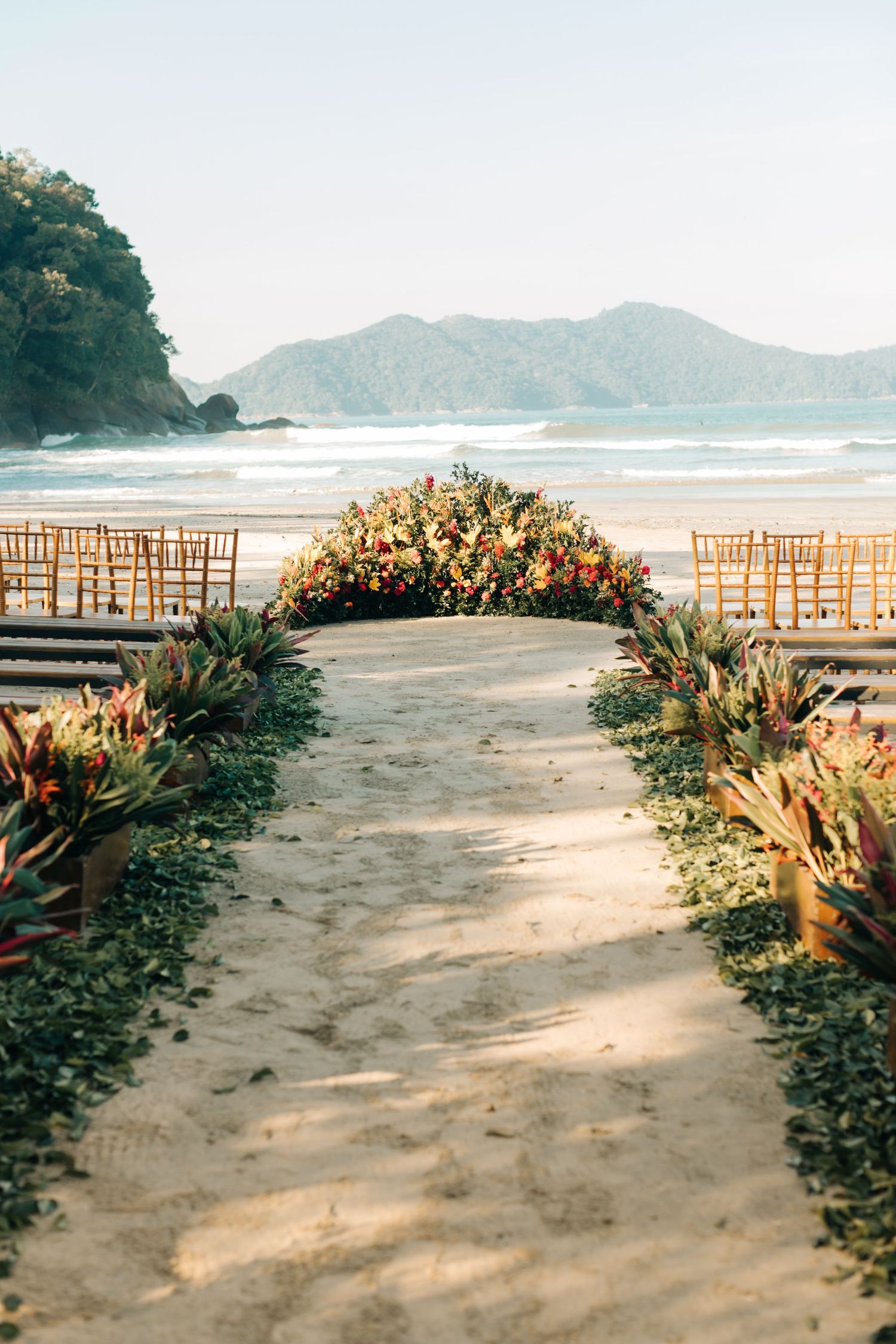Beachfront wedding ceremony setup with floral arch and wooden chairs facing the ocean.