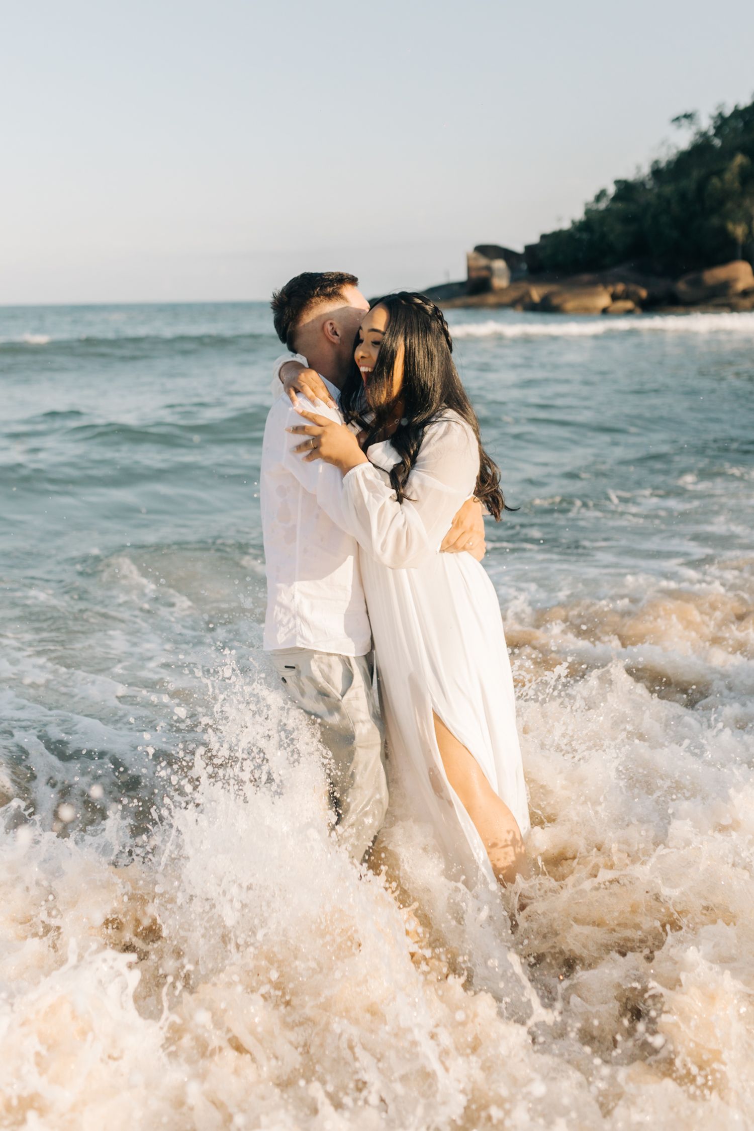 A couple plays joyfully in the ocean waves, splashing and embracing in white outfits at the beach during sunset.