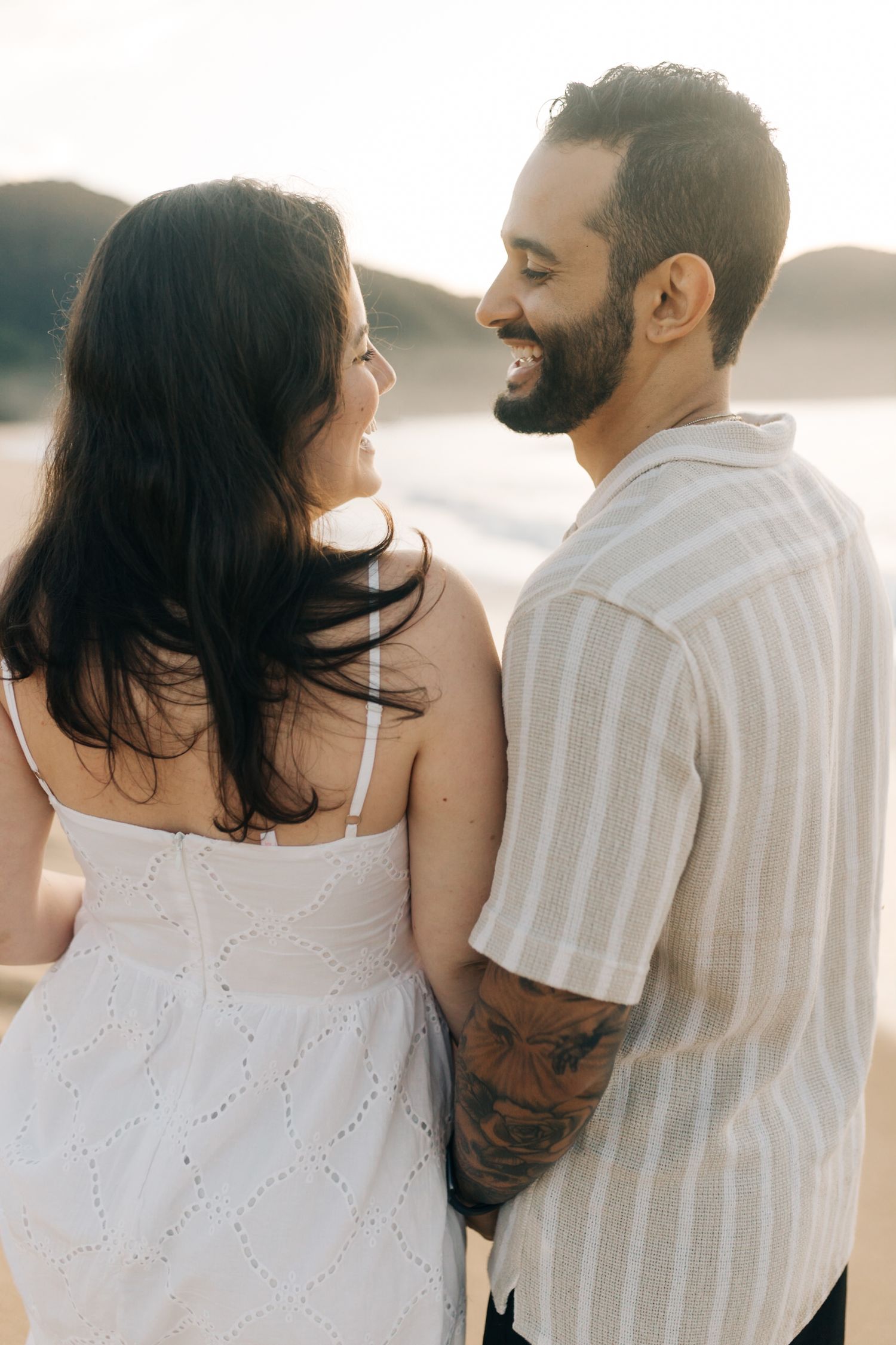 A romantic couple smiles at each other while enjoying a sunset beach moment.