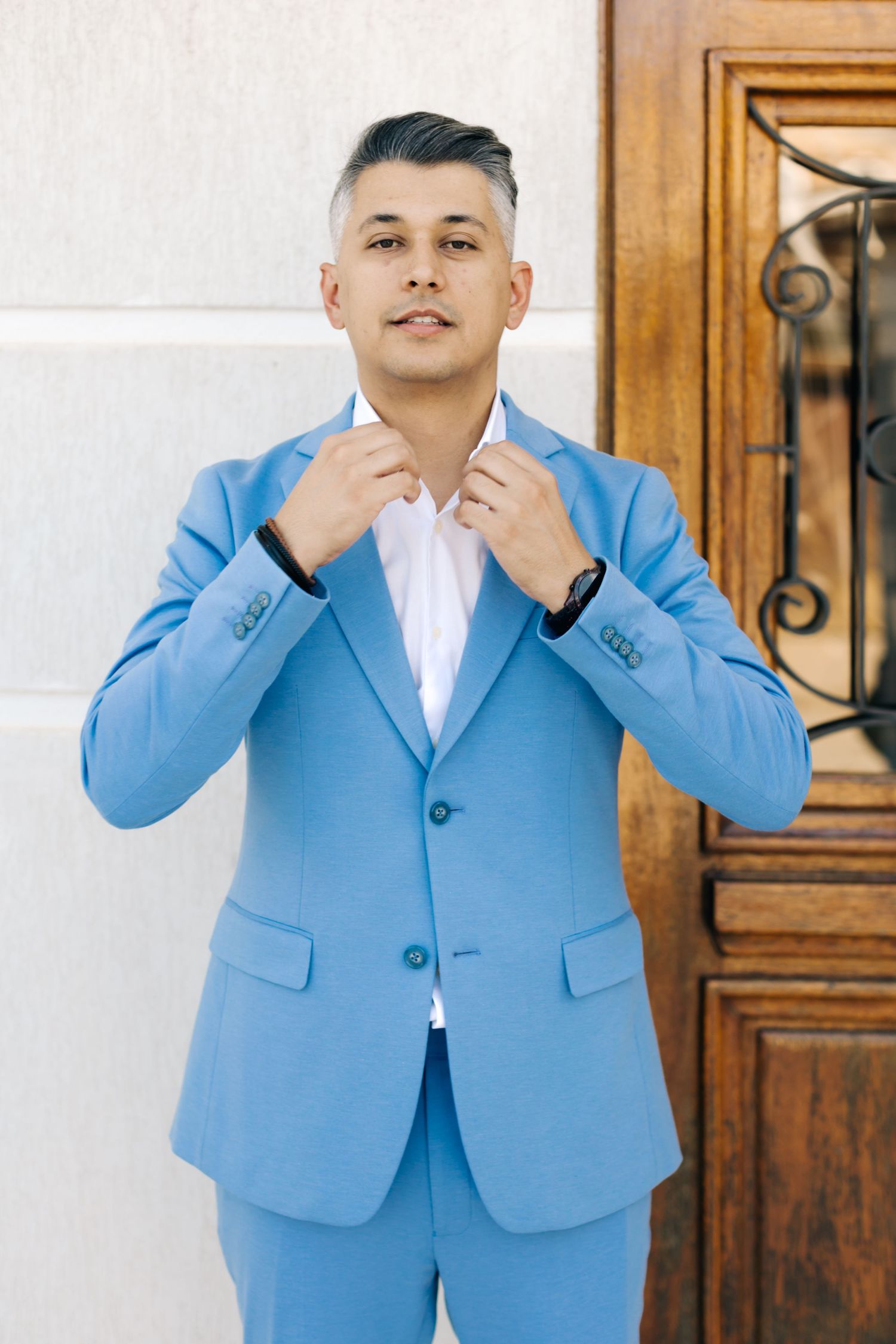 Person in light blue suit adjusting collar while standing against white wall with wooden door.