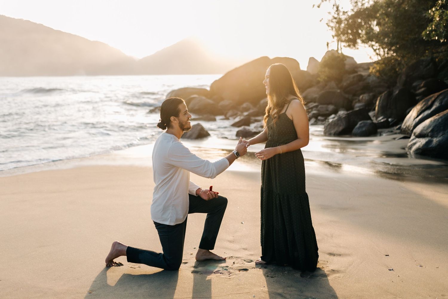 Romantic marriage proposal on beach with crashing waves and rocky coast at sunset.