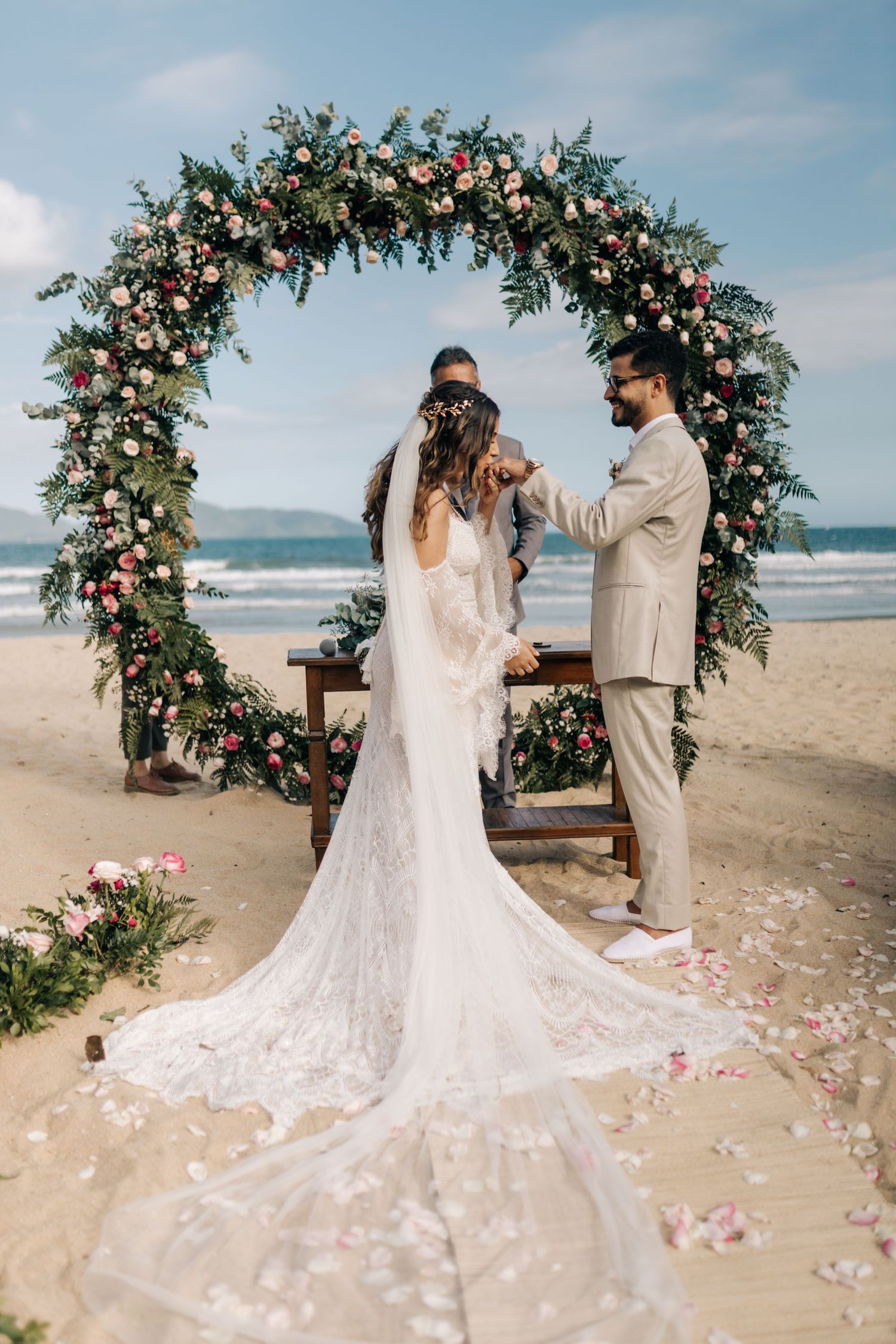Beautiful floral arch wedding ceremony on a beach with long flowing lace wedding dress train.