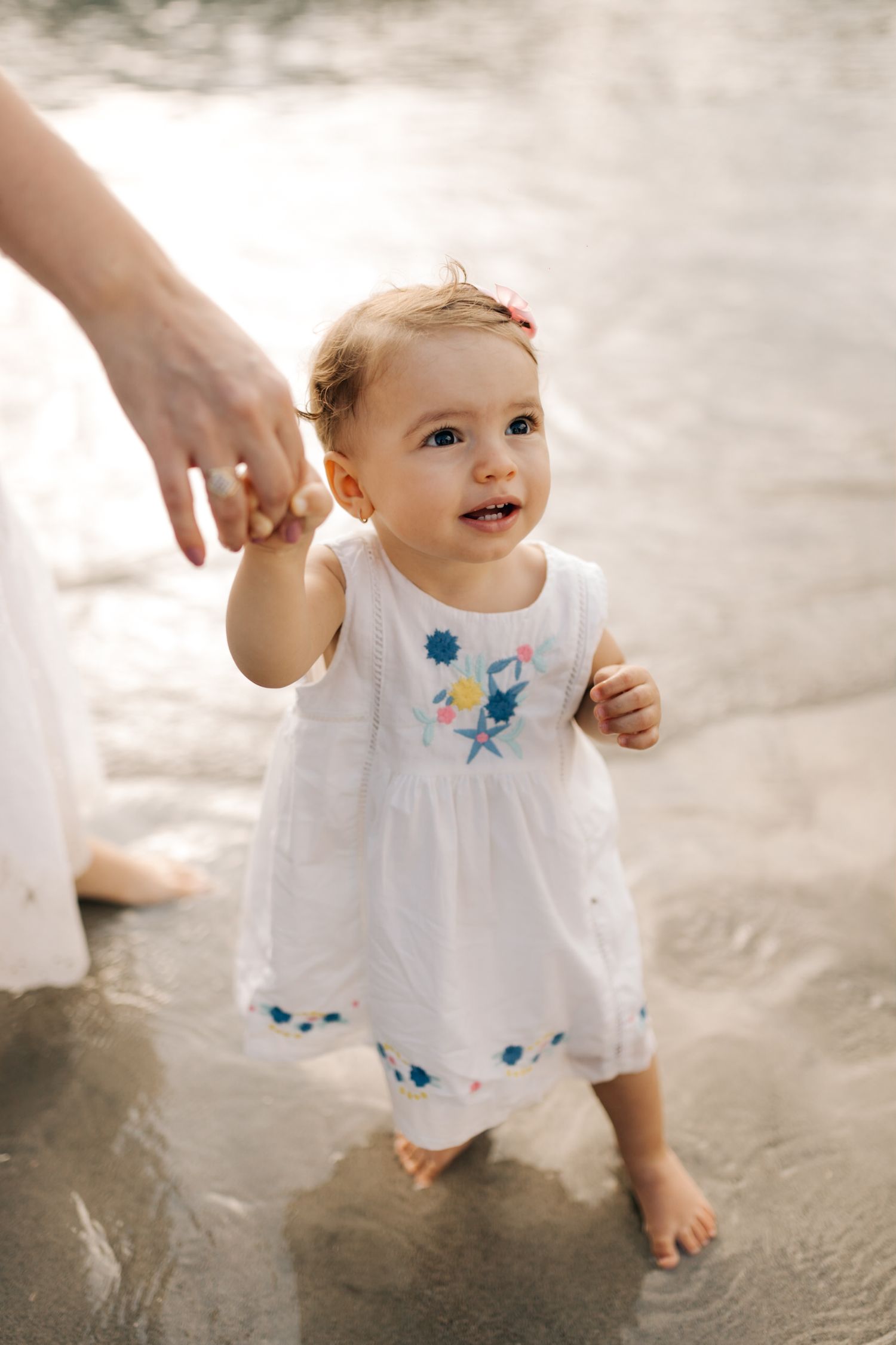 Toddler in white embroidered dress holding hand while walking in shallow water.