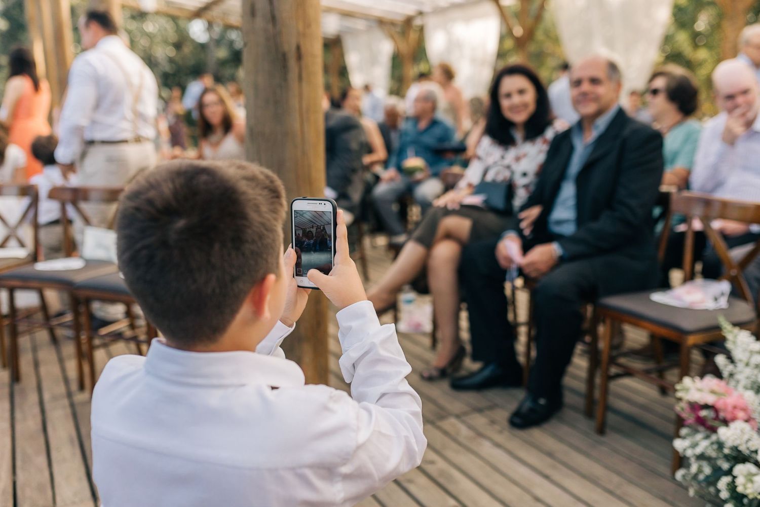 Young guest taking a photo with smartphone during an outdoor garden wedding ceremony.