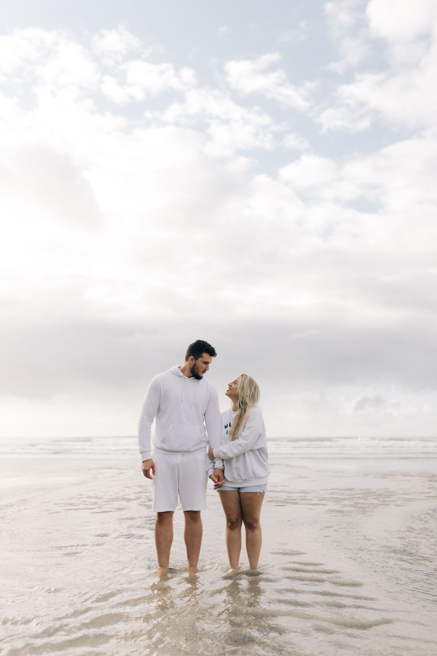 Couple in matching white outfits standing in shallow ocean water against cloudy sky.