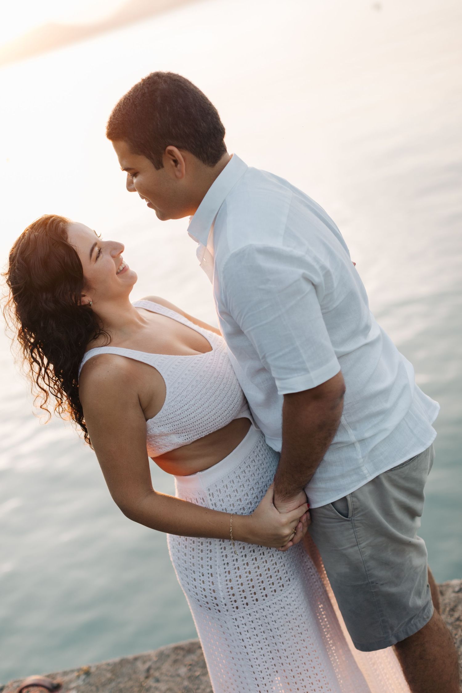 A couple in white outfits shares a tender embrace against a sunset water backdrop.