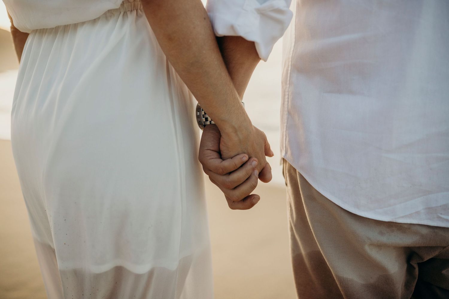 Close-up of couple holding hands on sandy beach during golden hour.