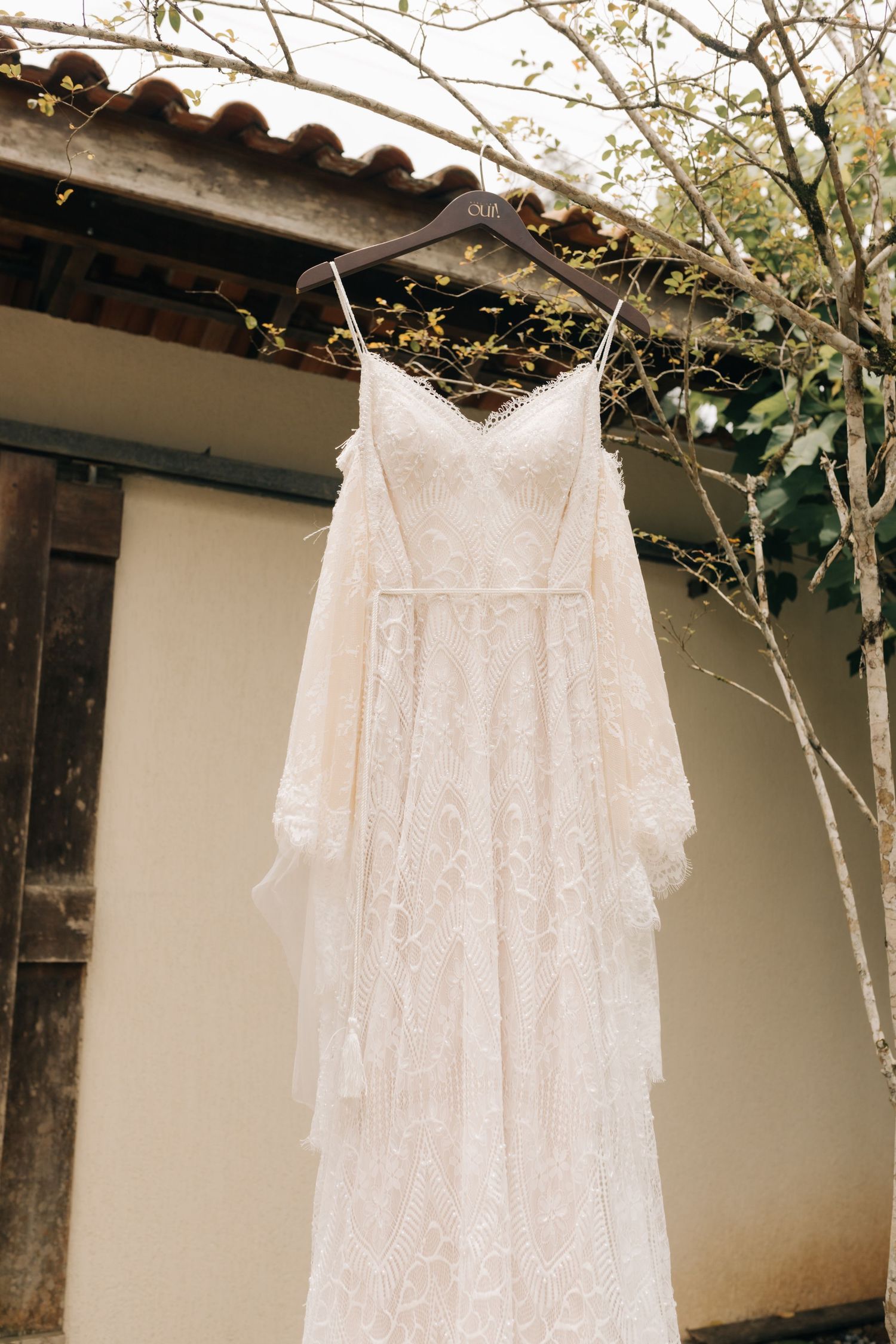 White lace wedding dress hanging outdoors against stucco wall with spring branches.