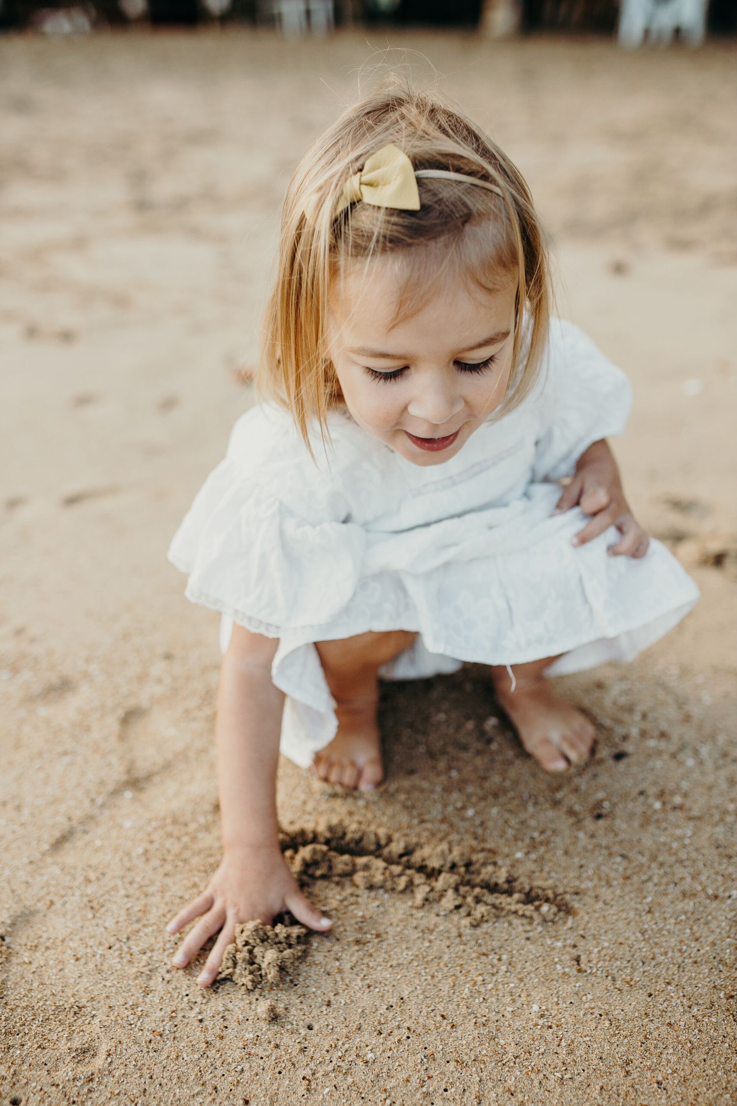A young child explores and plays in the beach sand wearing a white dress.