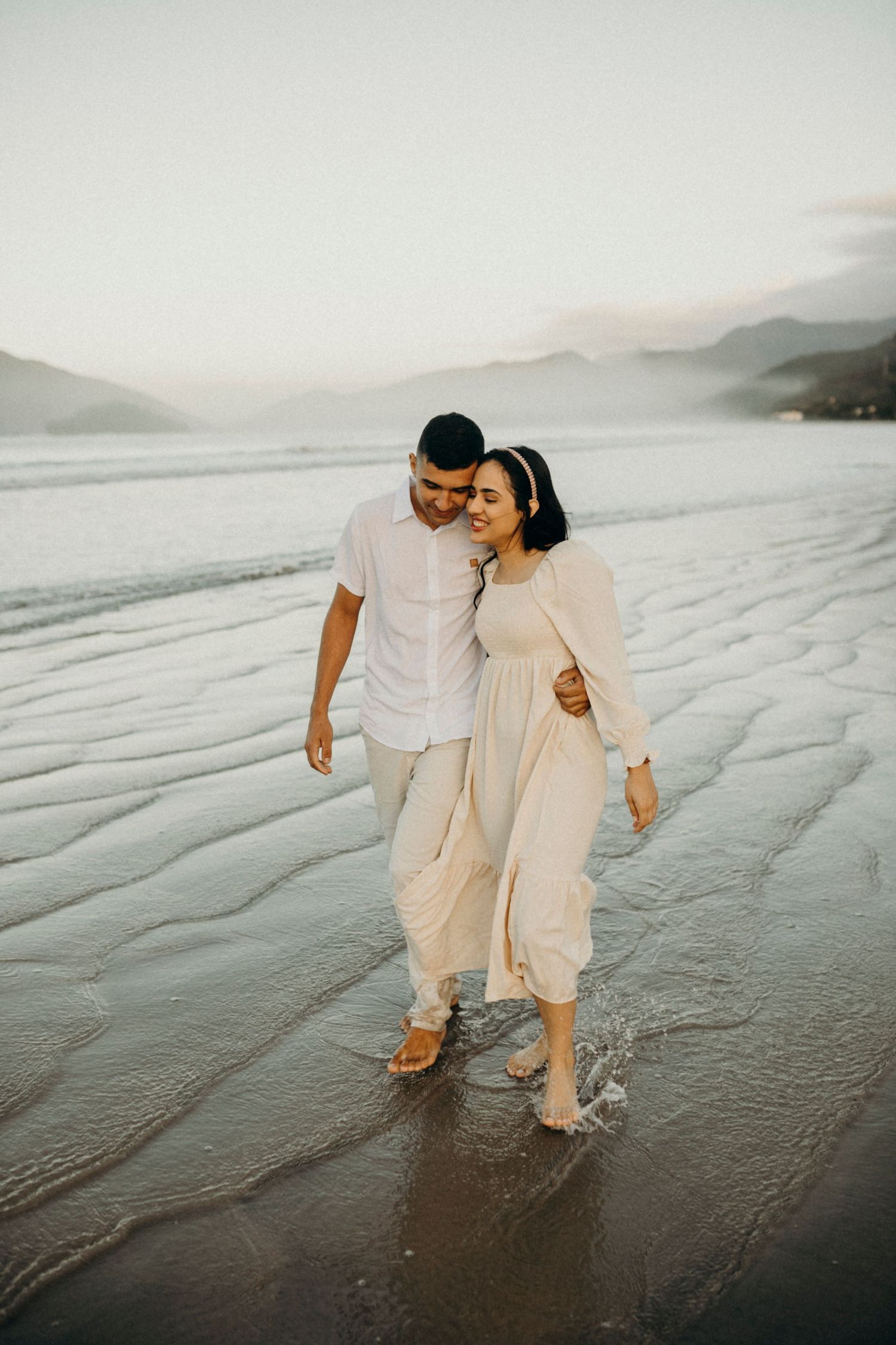 A couple dressed in white outfits shares a romantic moment walking along a misty beach at sunset.