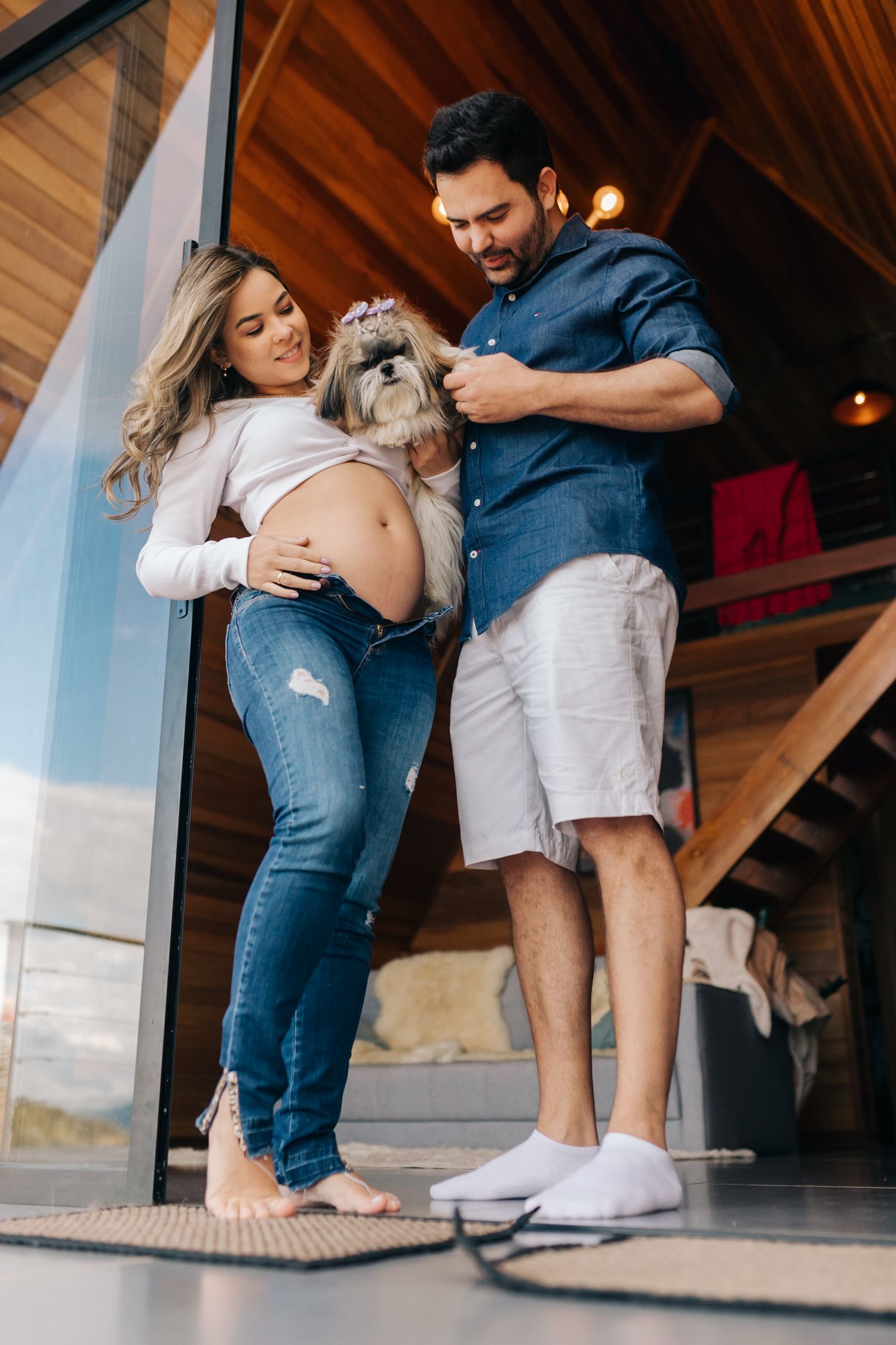An expecting couple poses with their small dog in a modern cabin setting.