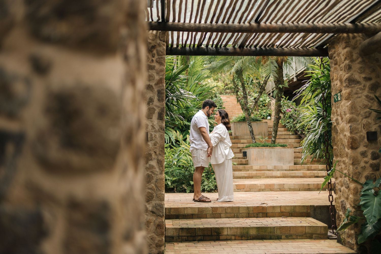 A couple in white outfits walks together through a garden pathway with stone columns and lush greenery.