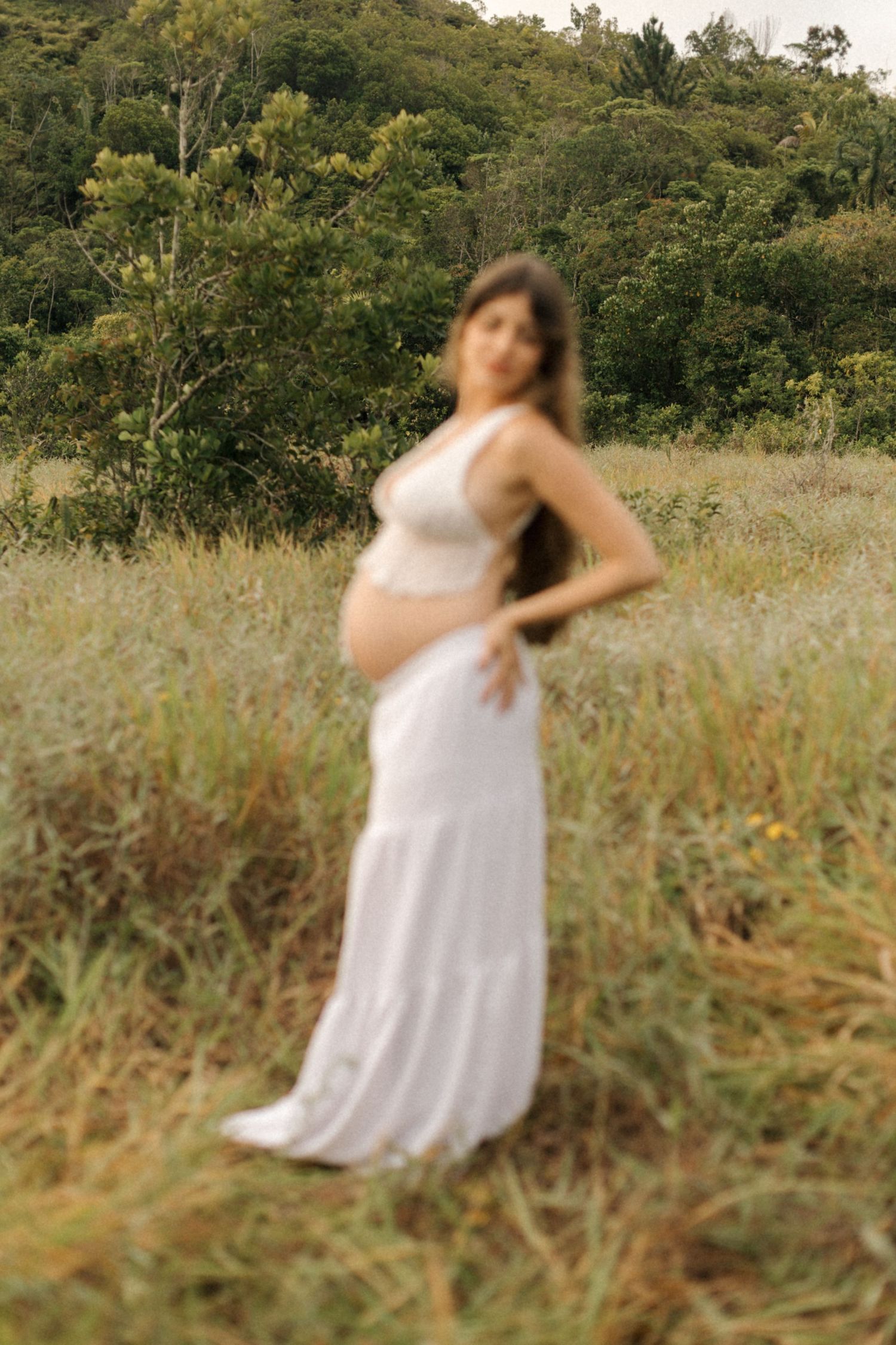 A white maternity gown flows elegantly in a natural meadow setting with lush green forest in the background.