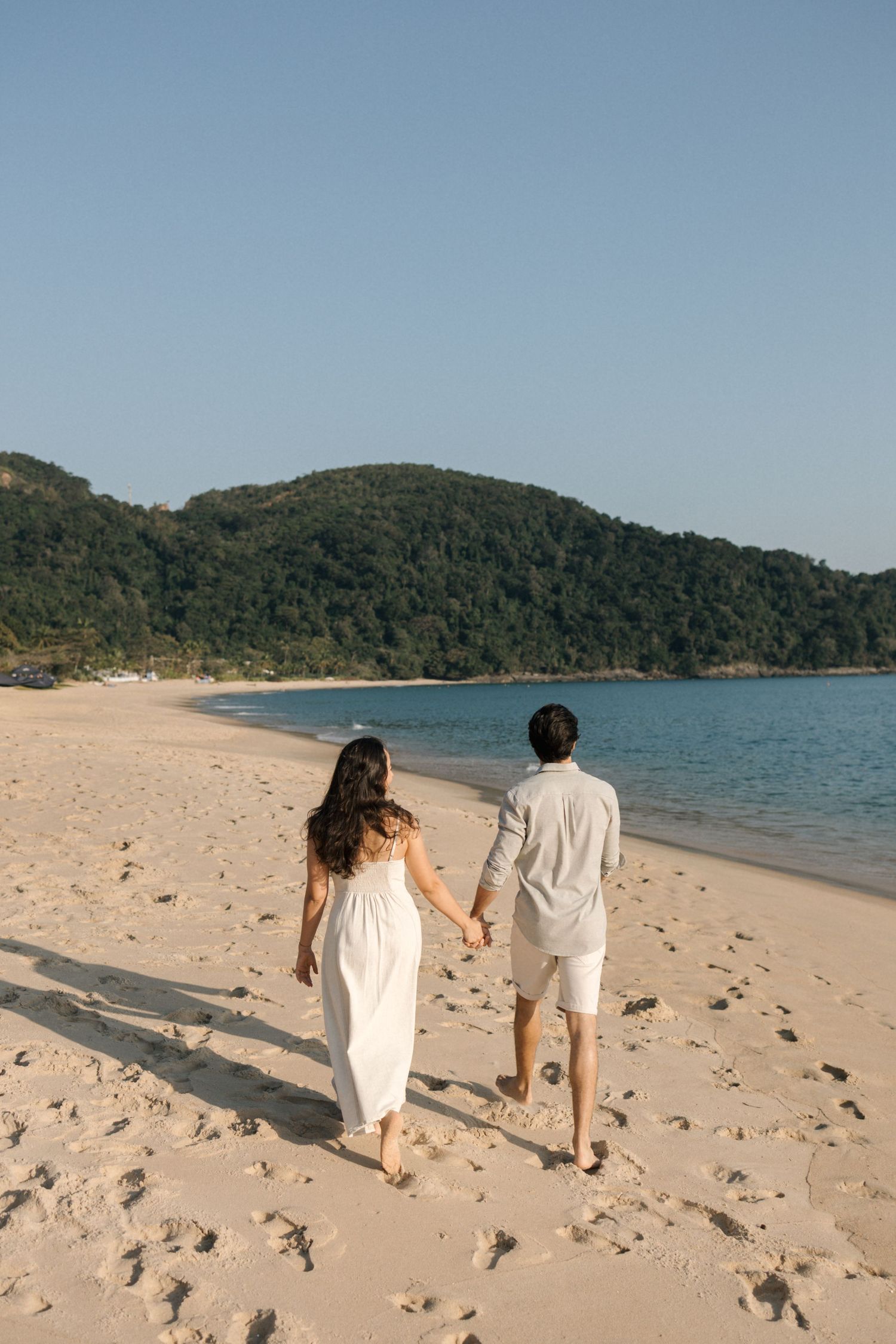 A couple in white outfits walking together along a scenic beach with turquoise water, mountains and blue sky.