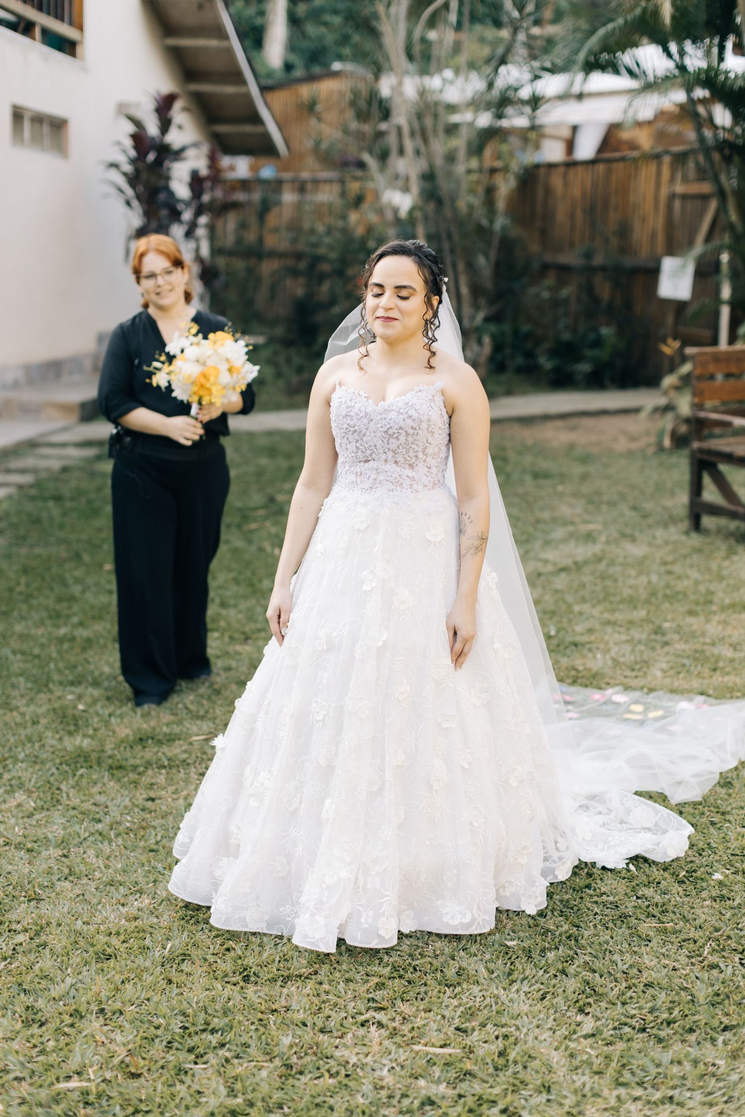 Bride in white lace wedding gown poses on grass while attendant holds flowers in background.