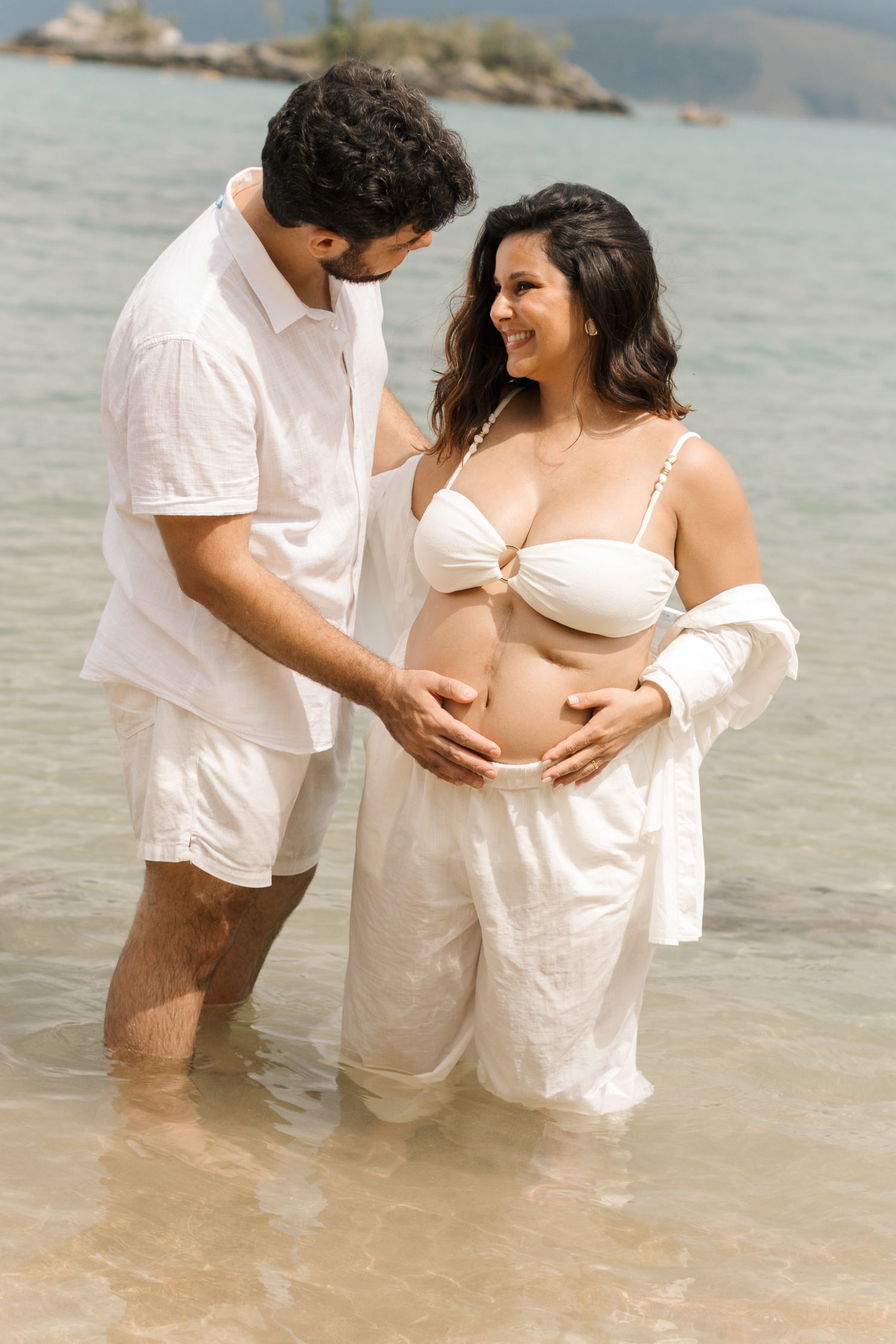 Expecting couple shares intimate moment while standing in shallow ocean water wearing white outfits at beach.