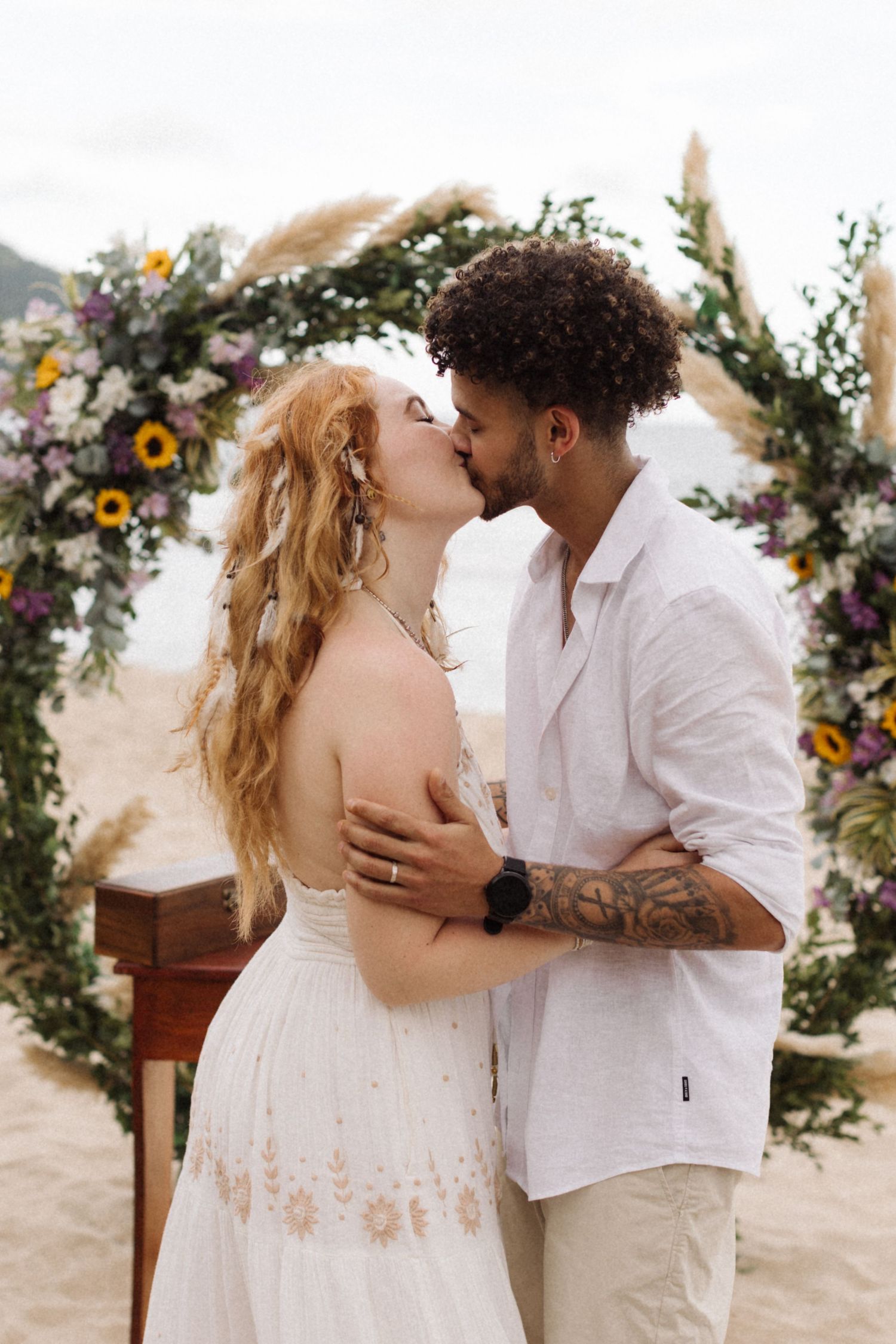 Romantic beach wedding kiss under a floral arch decorated with sunflowers and wildflowers.