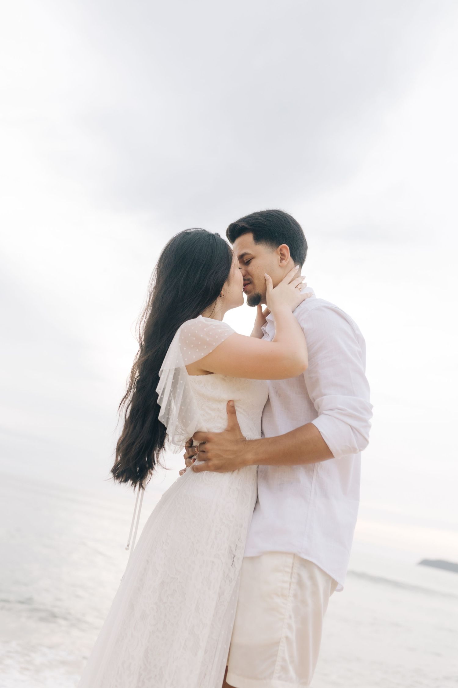 Couple in white outfits sharing intimate moments against cloudy beach backdrop.