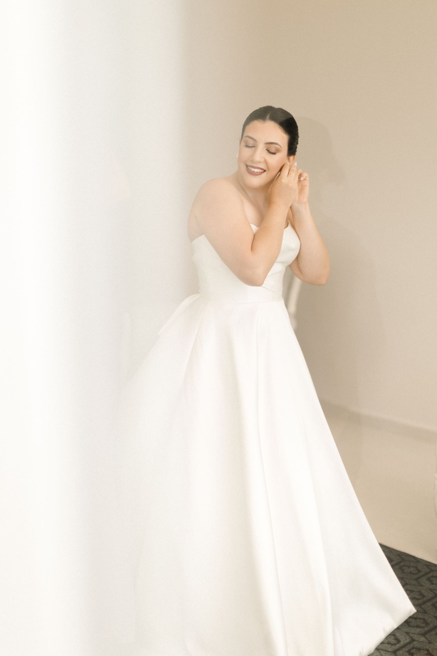 Person wearing elegant white wedding gown poses against light wall in bridal portrait.