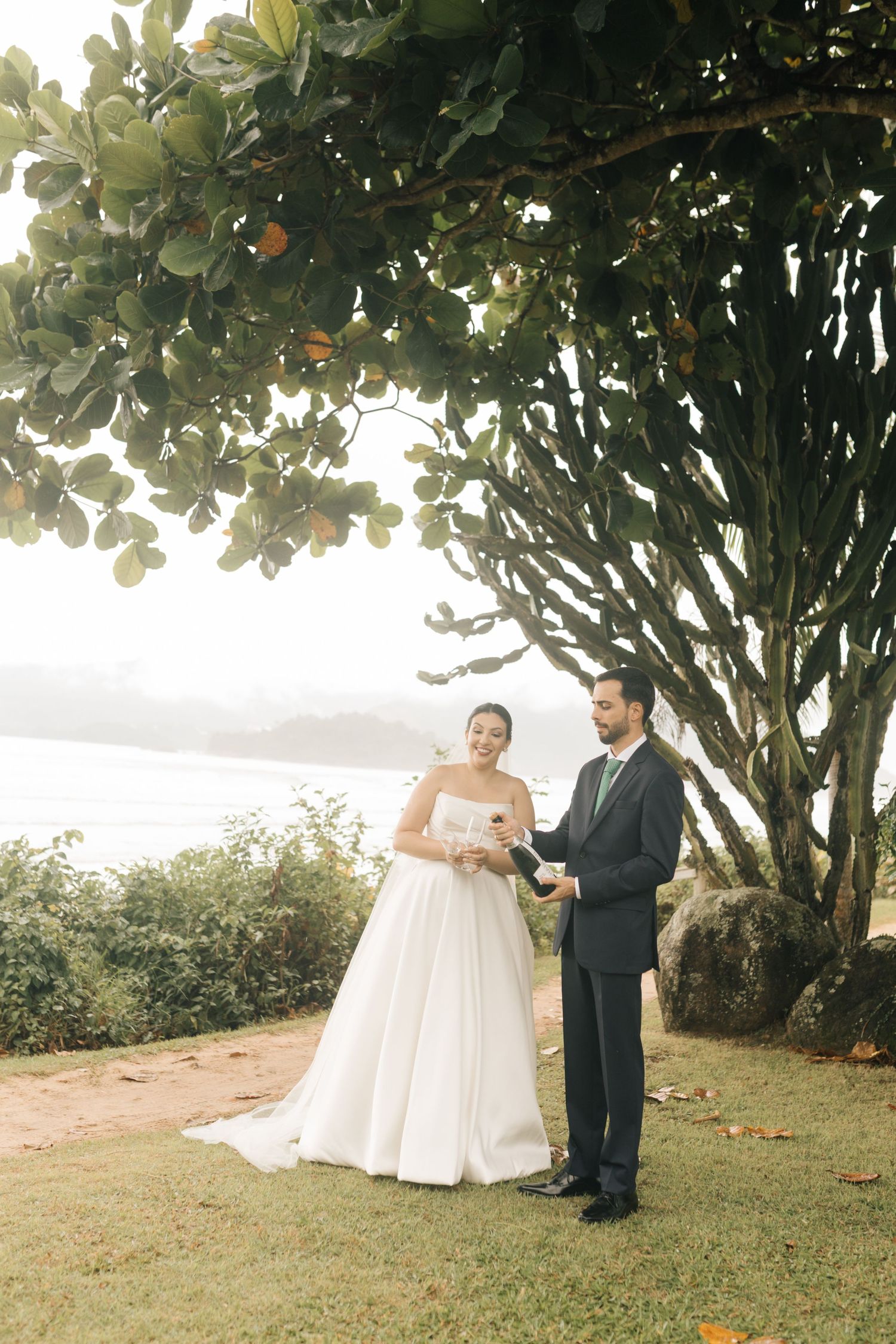 Wedding couple stands under a large tree overlooking a scenic coastal view.