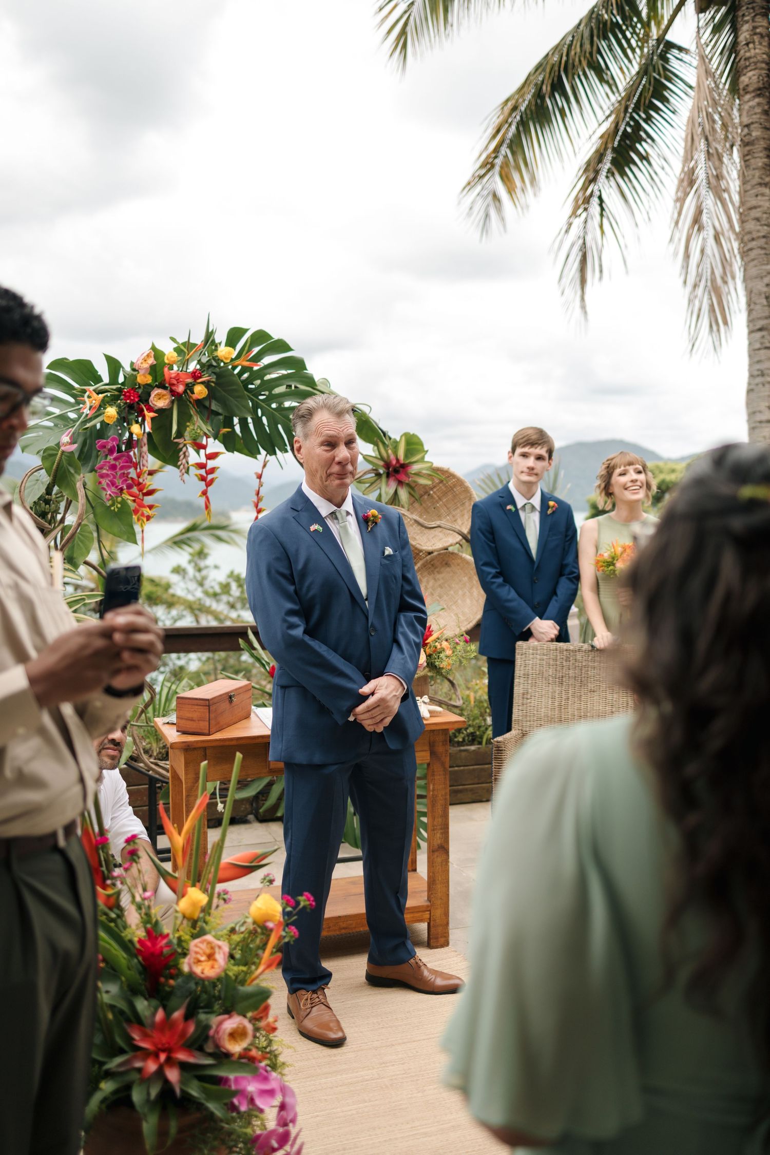Wedding ceremony guests in formal attire gather around tropical floral arrangements outdoors.