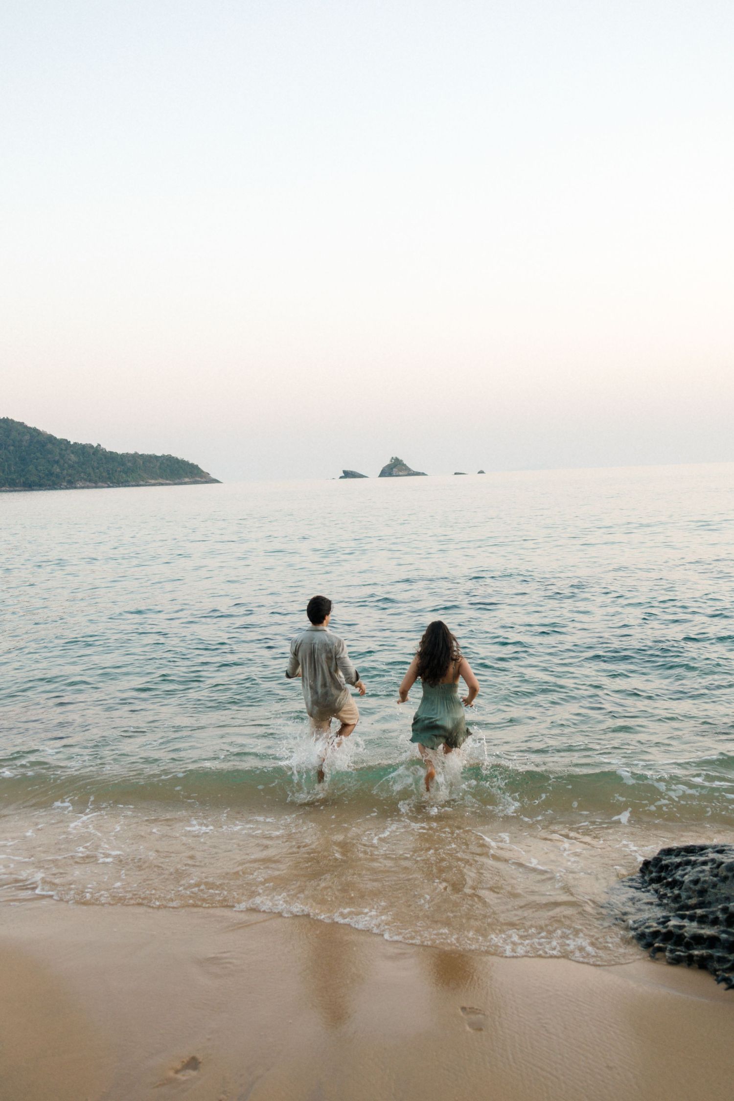 Two people sit at the water's edge watching waves roll in during sunset at the beach.