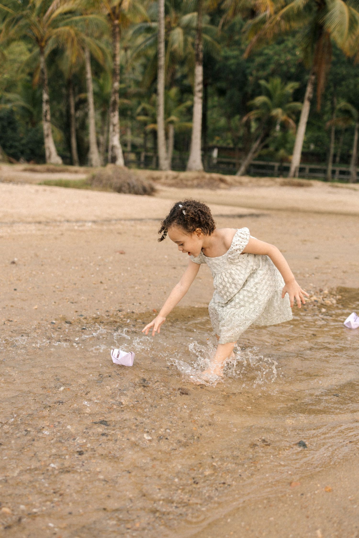 A child in a white dress splashes and plays in shallow beach water near palm trees on a tropical shore.