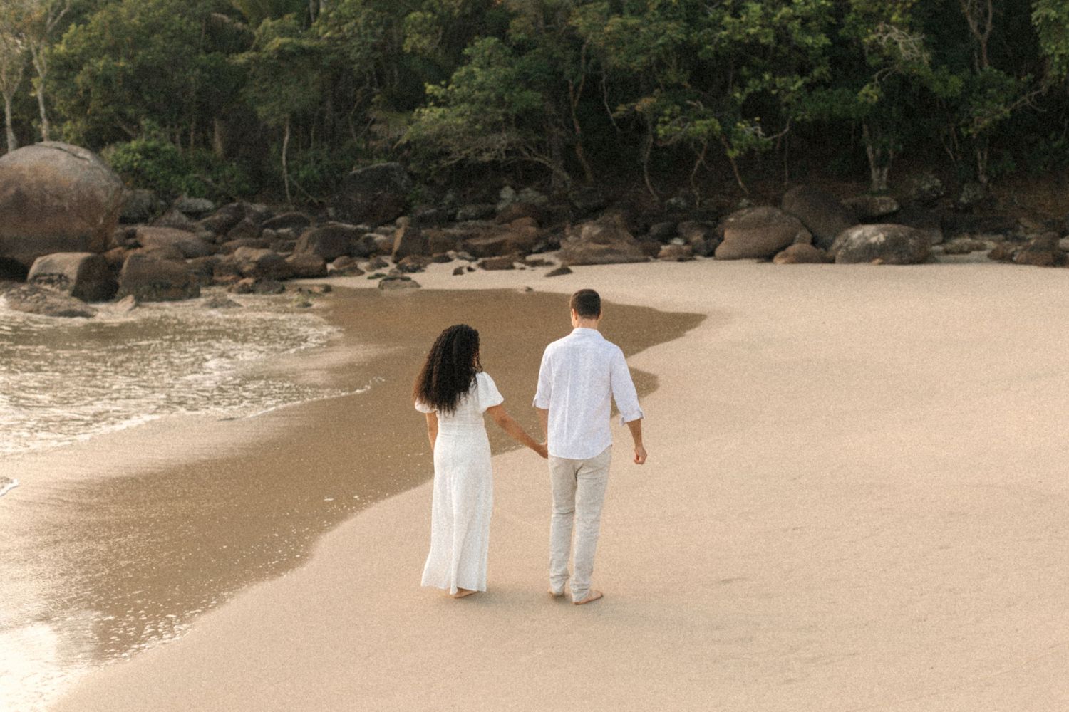 A couple wearing white clothing walks hand in hand along a secluded tropical beach surrounded by rocks and trees.