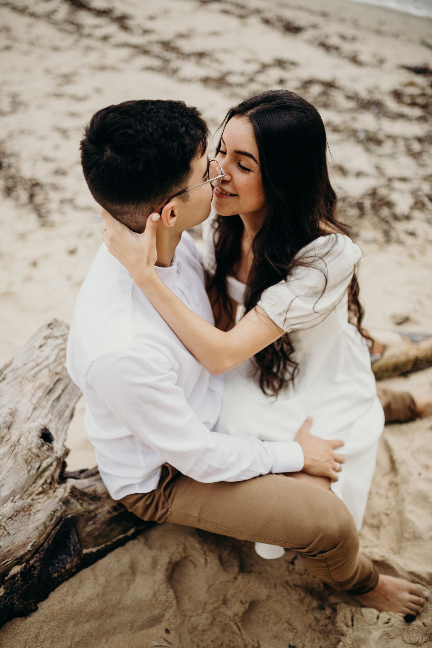 Couple shares romantic moment on beach with driftwood and sand.