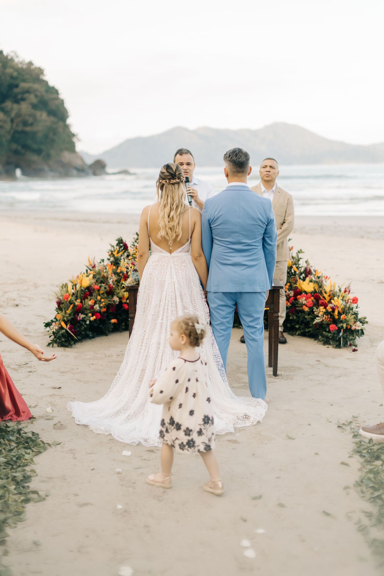 Couple walking barefoot on sandy beach with vibrant floral arrangements and mountain views.