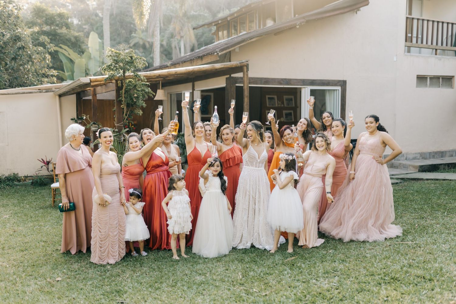 Bridal party in shades of pink and red gathered on grass outside venue.