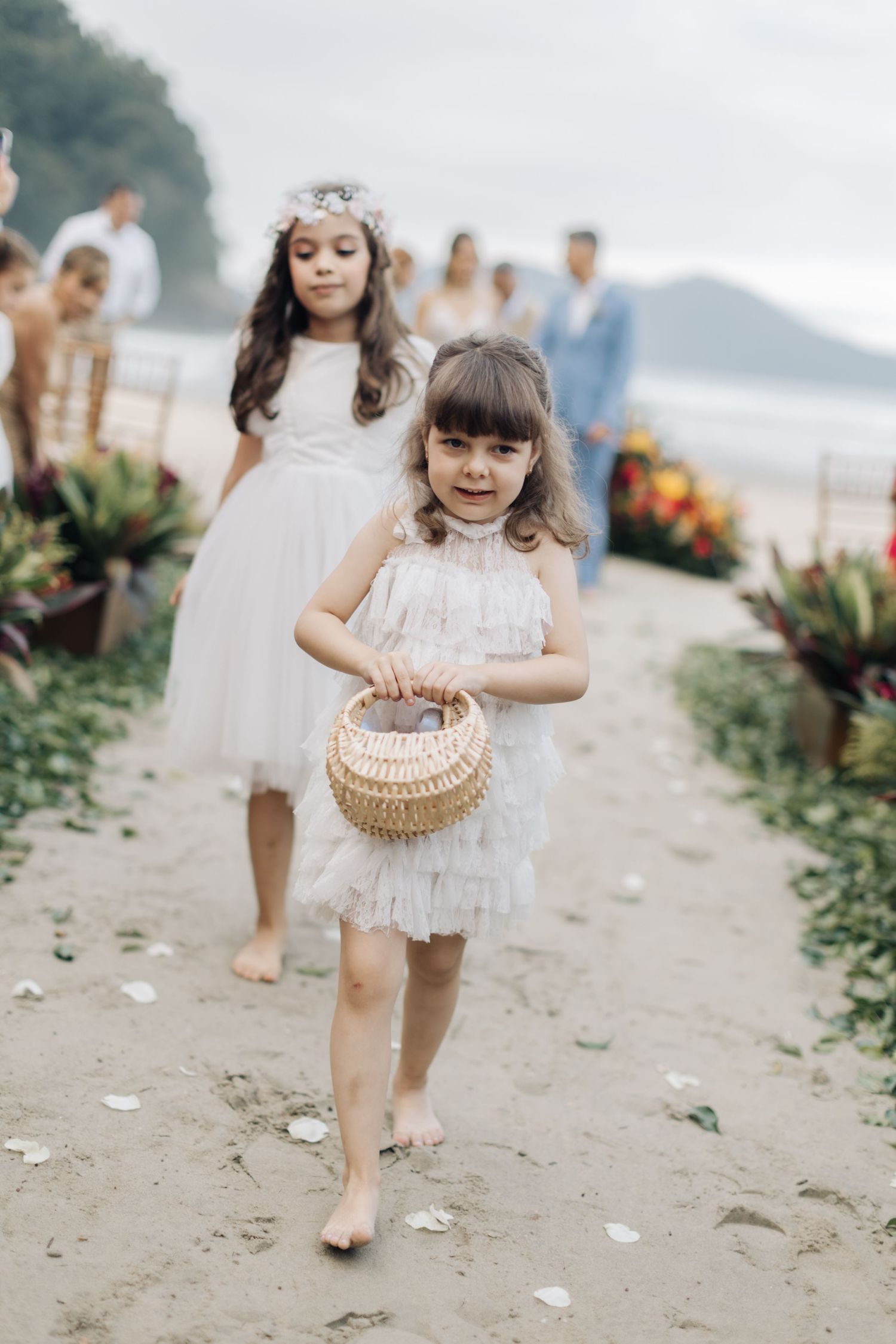 Young flower girl in a white lace dress carrying a woven basket on a sandy beach.