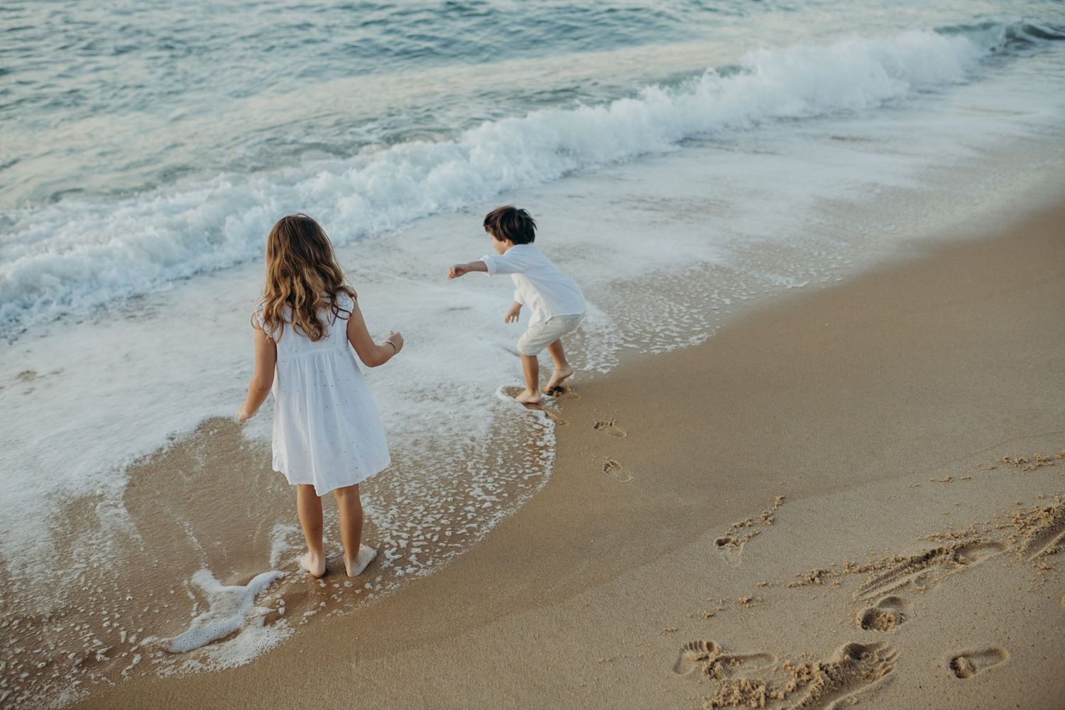 Beachgoers in light clothing playing near the ocean waves as they crash on shore.