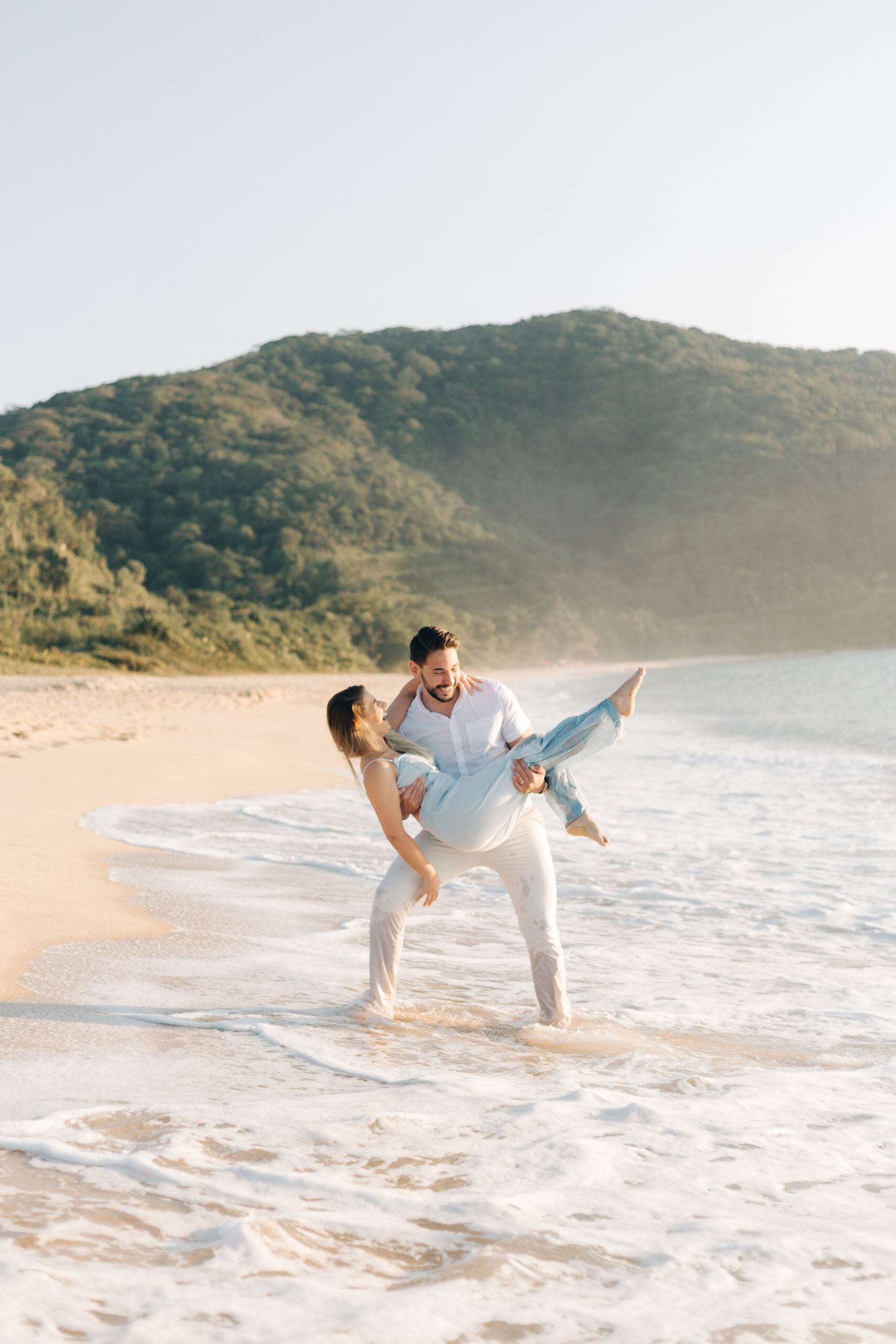 Playful couple dancing in ocean waves on scenic beach with mountains in background.