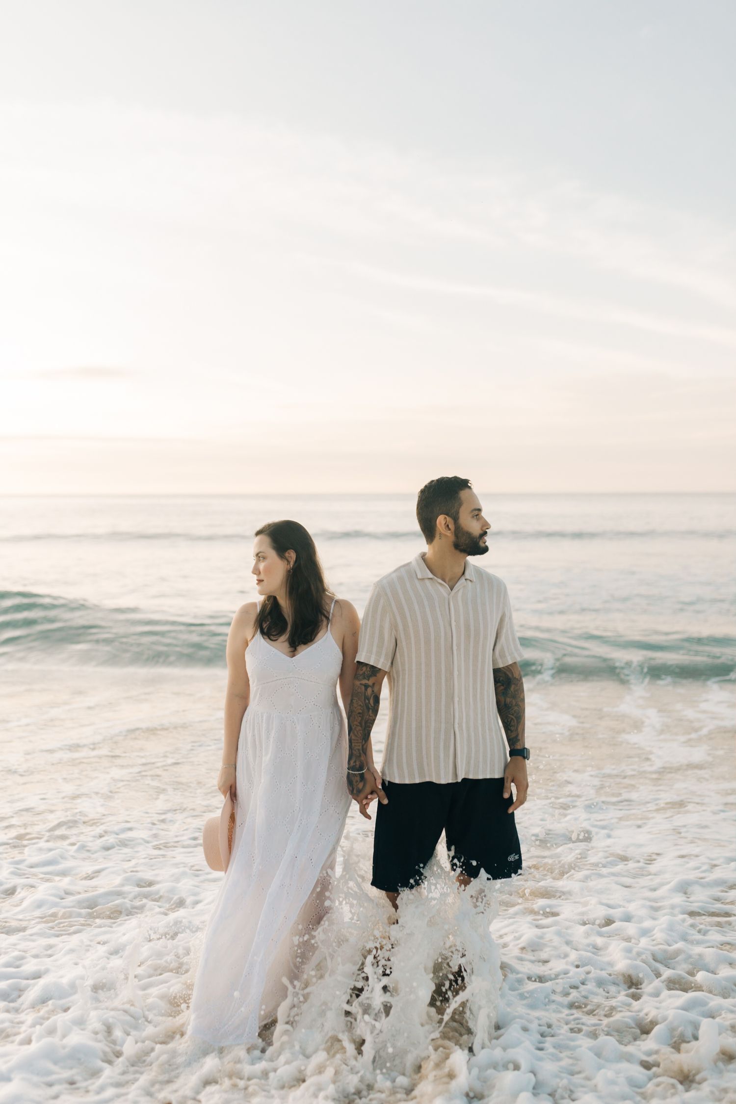 Couple stands in ocean waves together wearing white and casual beach attire.