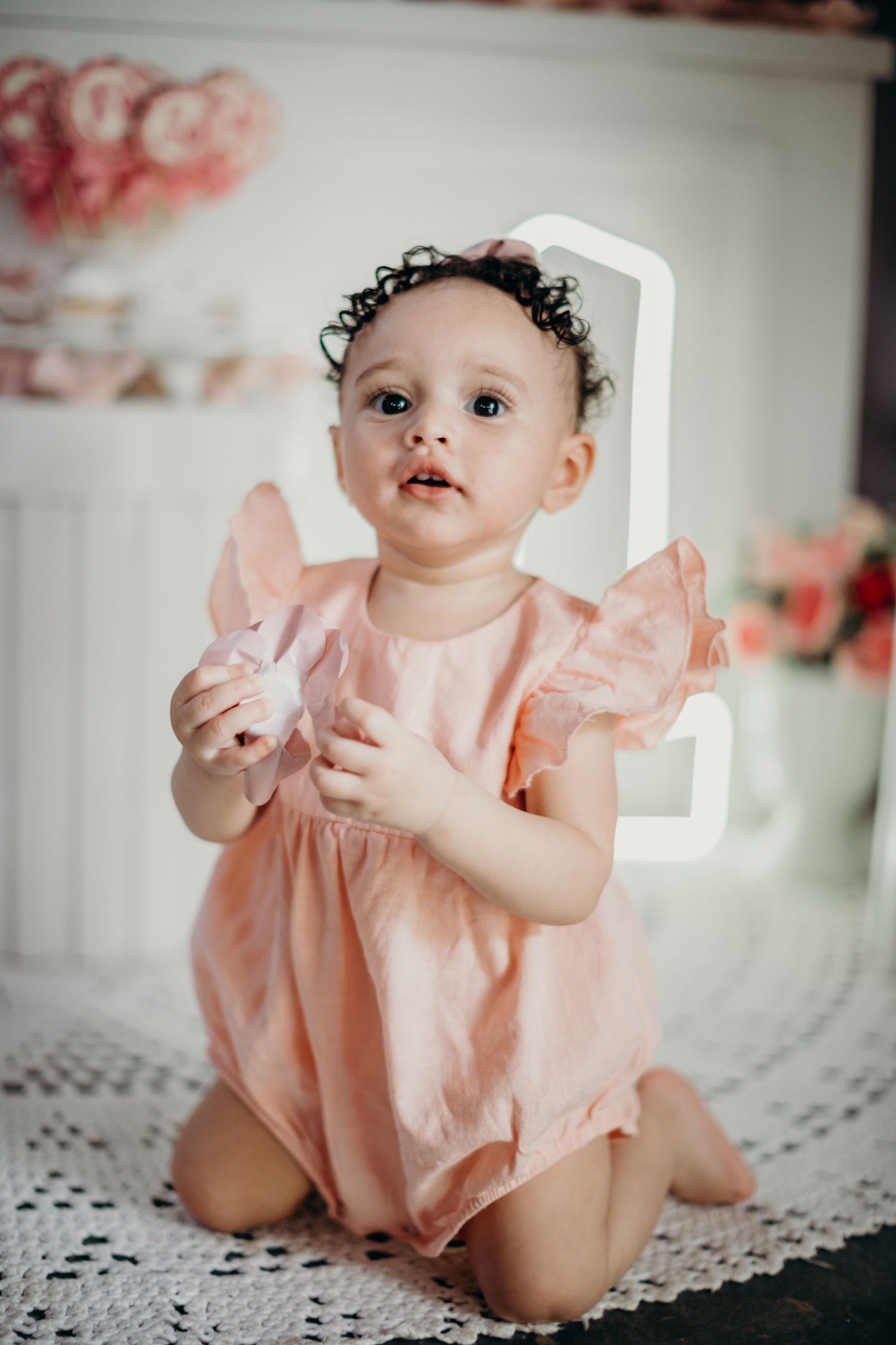Baby in pink ruffled outfit posing on white lace surface during celebration.