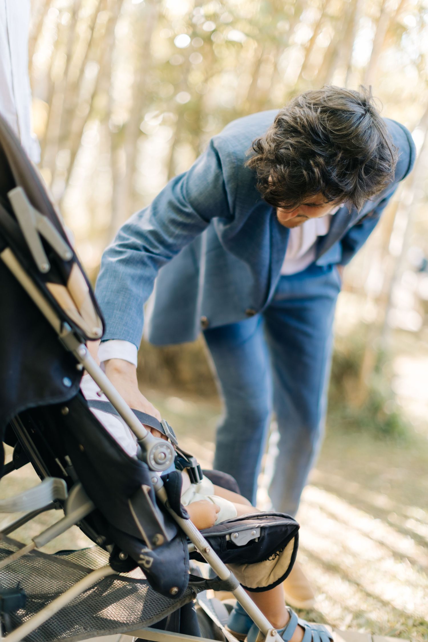 Person in blue jacket leaning over to adjust a baby stroller in a sunlit wooded area.