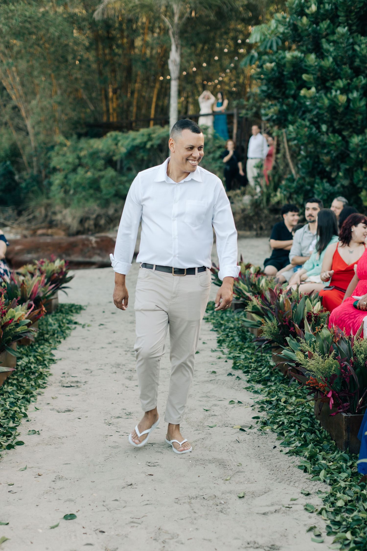 Person in white shirt and khaki pants walks down sandy aisle at outdoor ceremony.