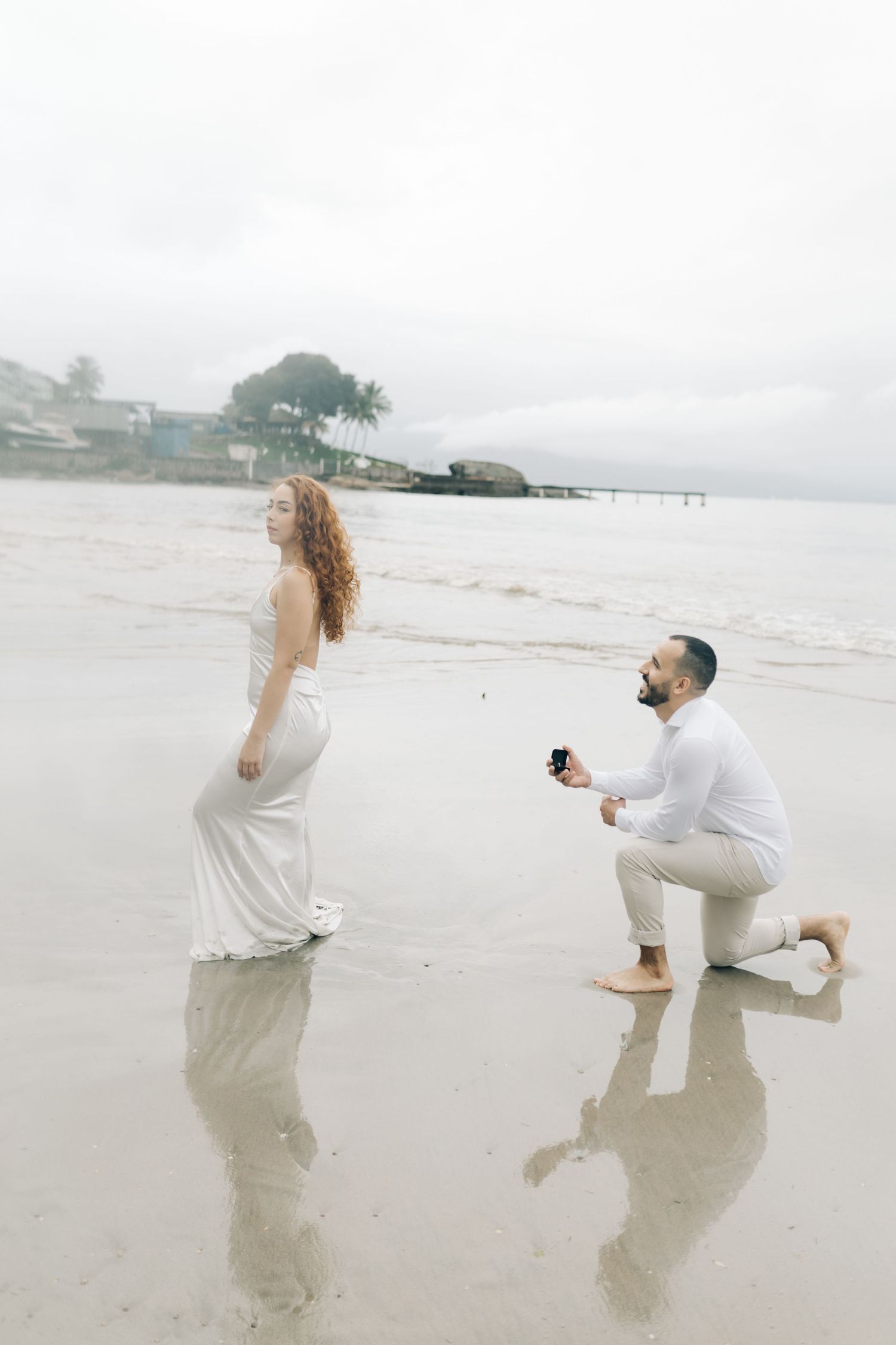 Marriage proposal scene on misty beach with couple in white outfits.