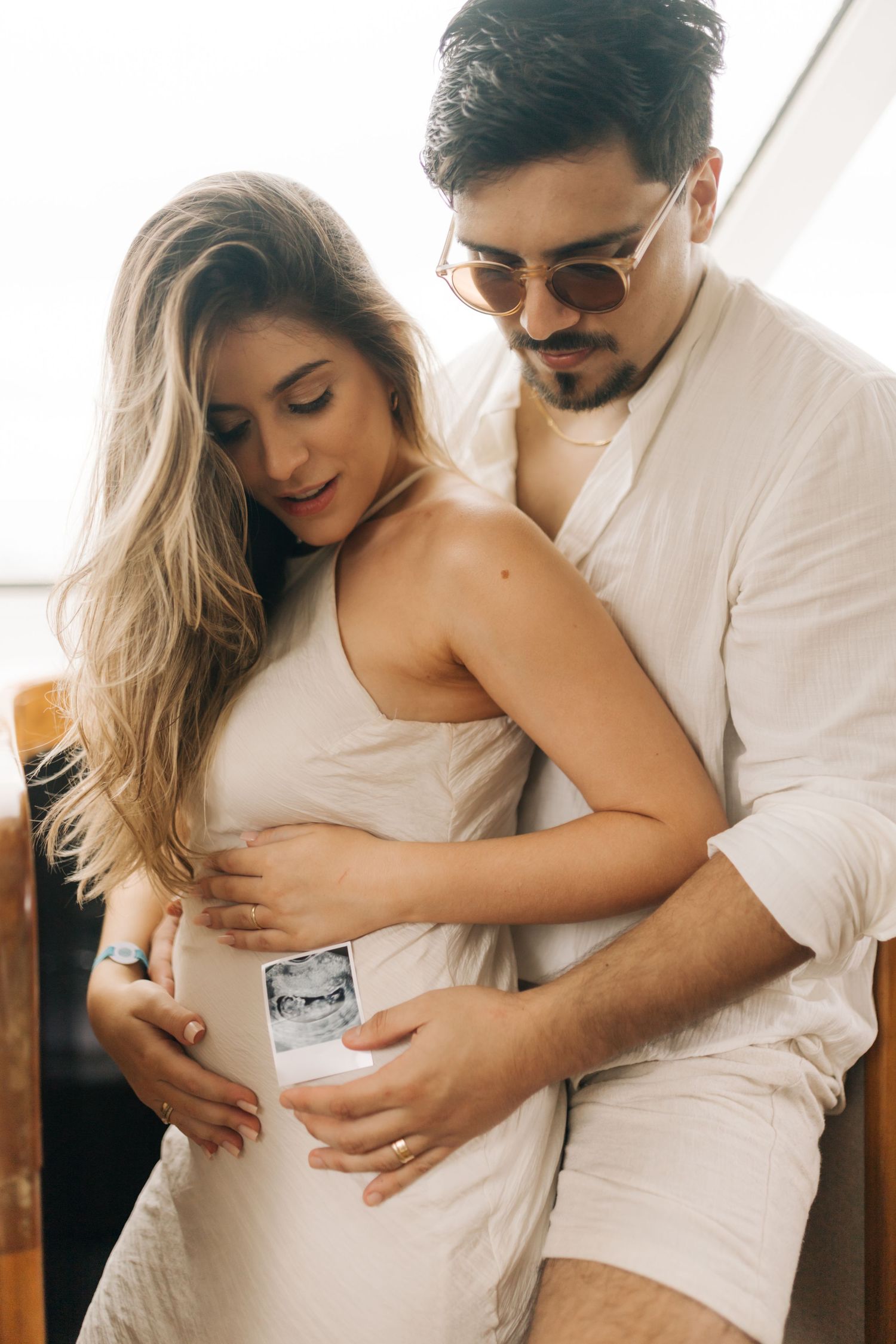 Couple holding an ultrasound photo while embracing in casual white outfits.