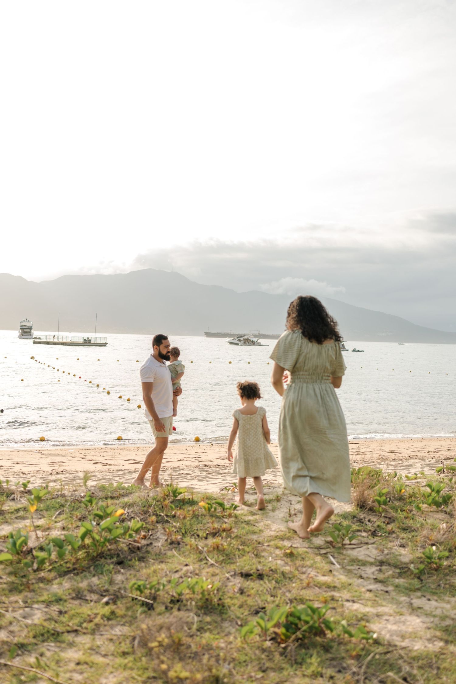 Family walking together along a scenic beach with mountains and boats visible in the distance.
