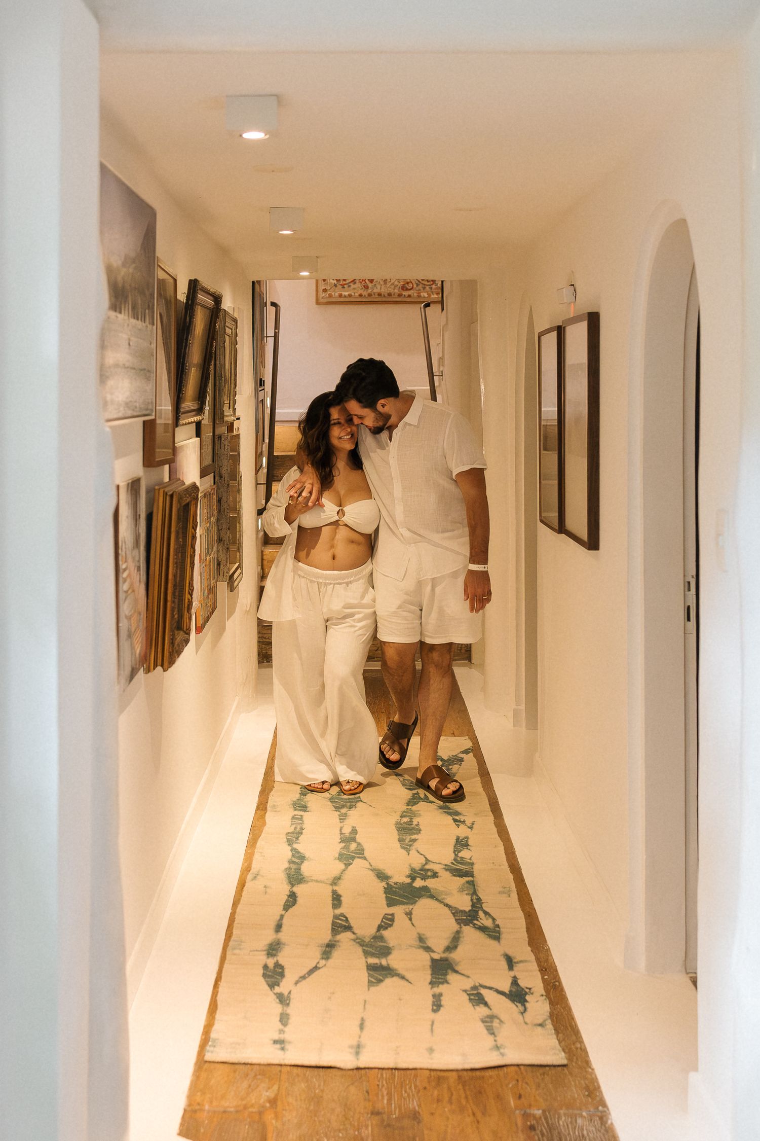 A couple walks together down a narrow hallway with white walls and a patterned runner rug.