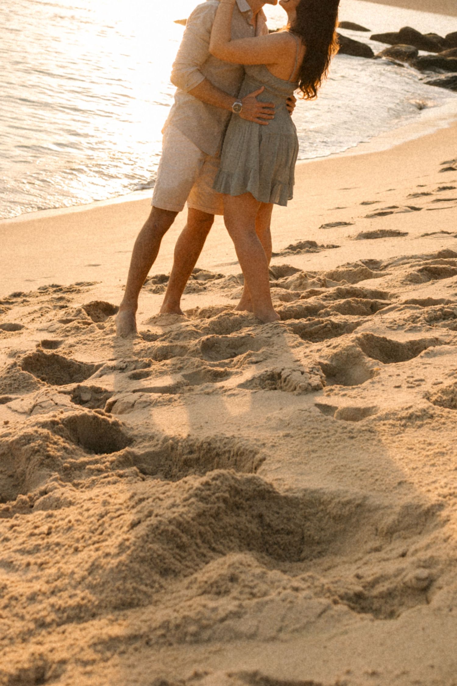 Two people stand close together on a sandy beach at sunset casting long shadows.