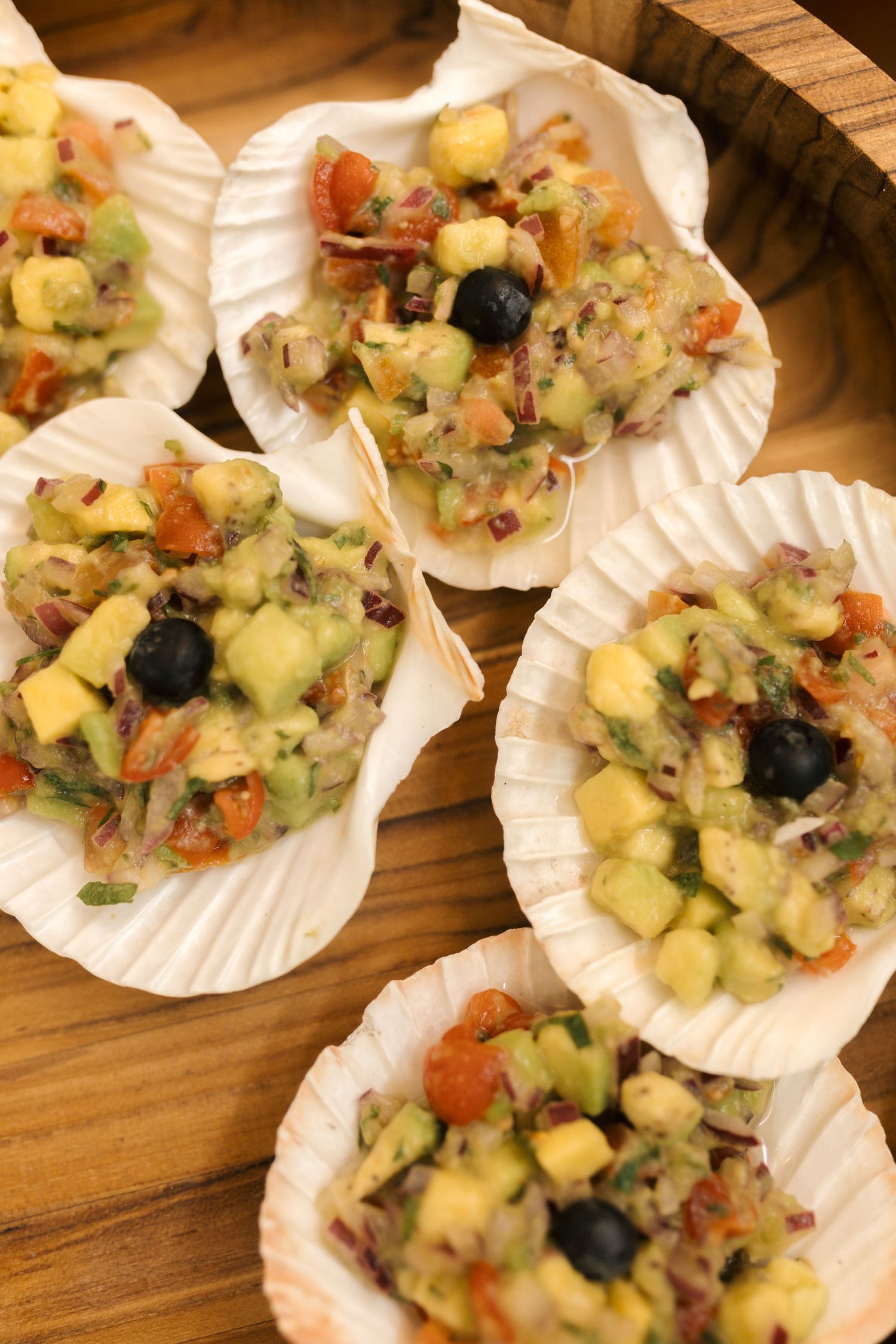 Seafood appetizers served in scallop shells with colorful vegetables and olives on a wooden surface.