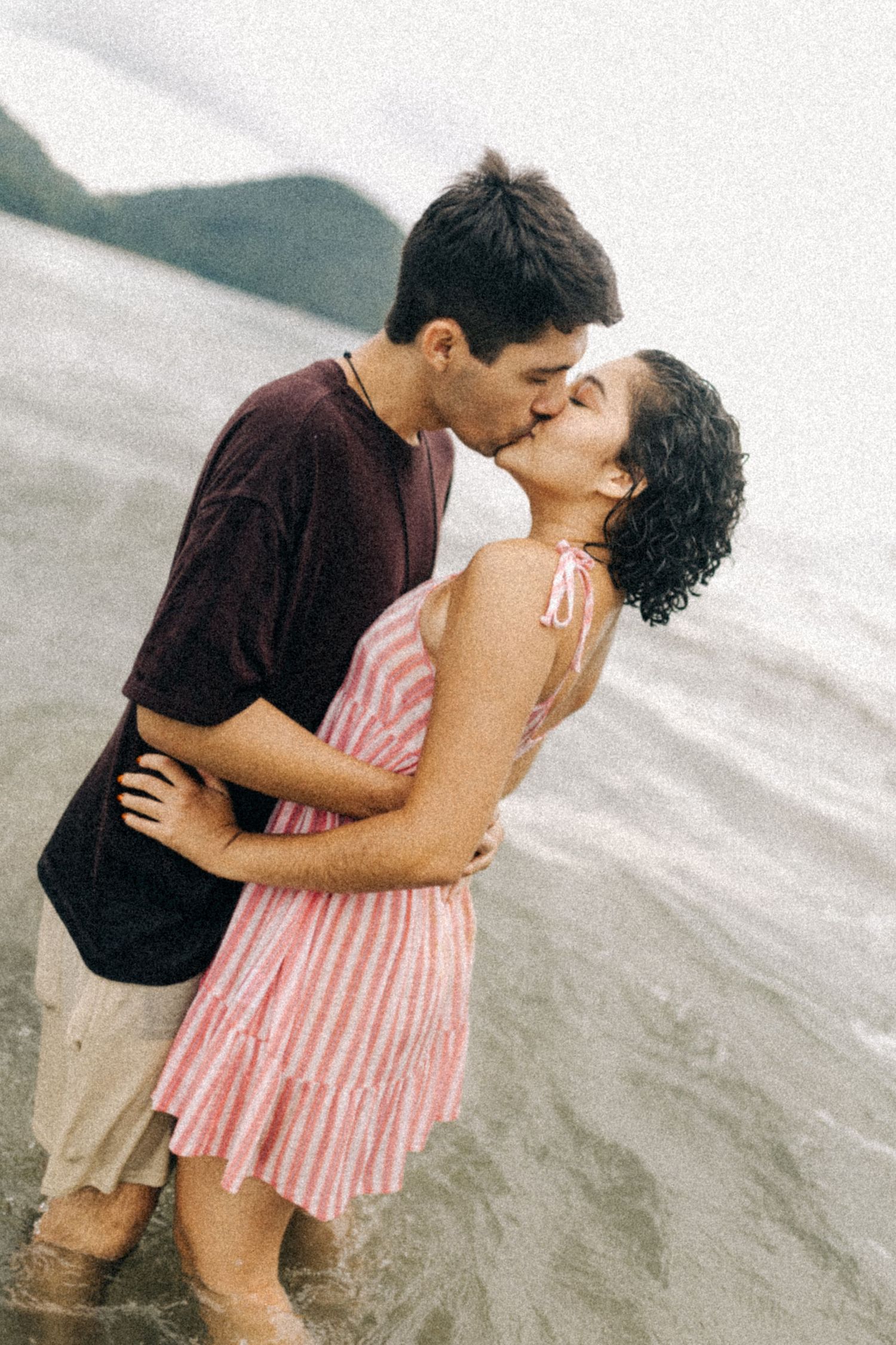 Um casal dá um beijo romântico na praia, com a mulher com vestido listrado rosa e o homem com camisa bordô.