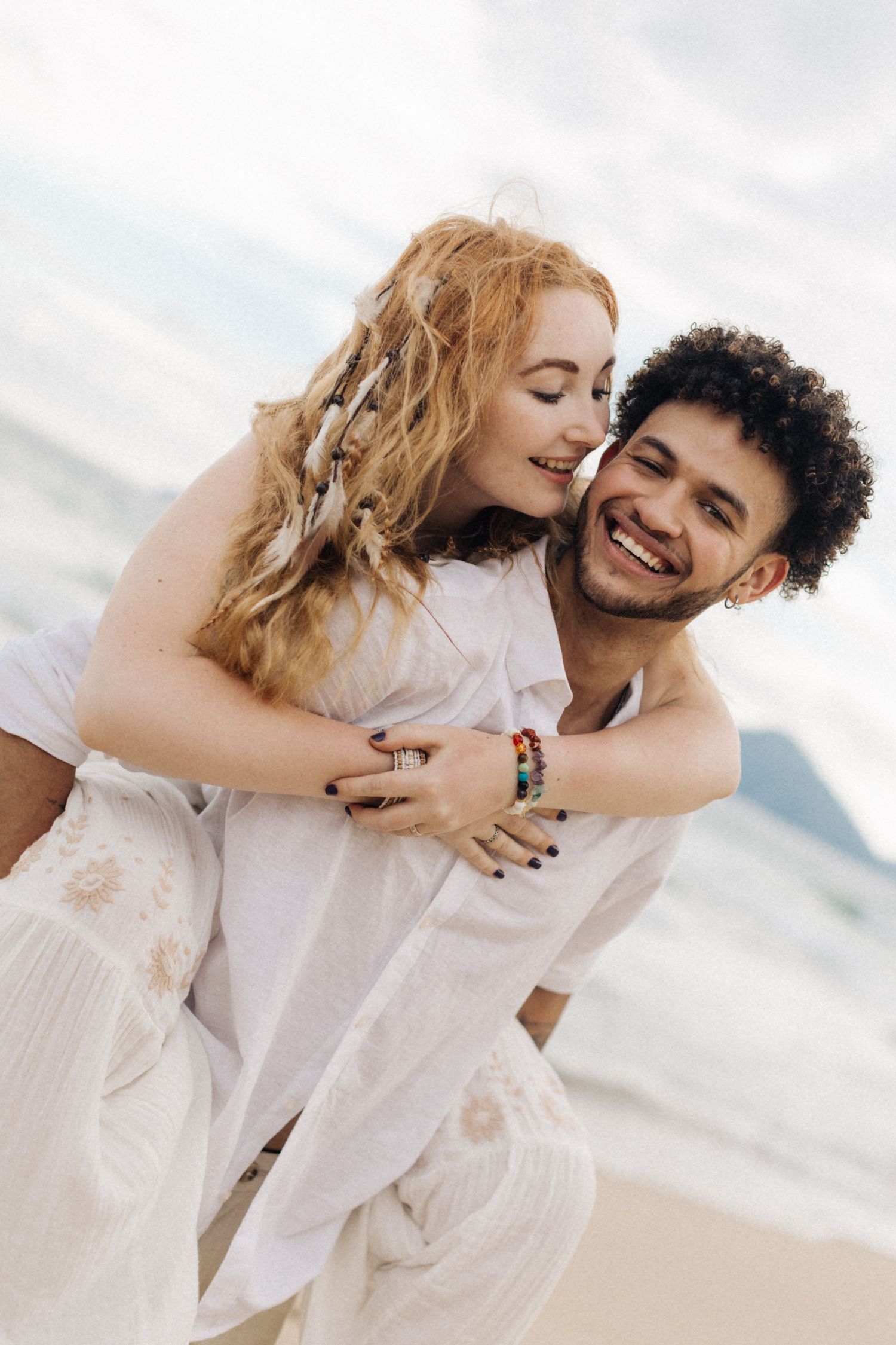 A couple in white outfits shares joyful moments together during a beach photoshoot.