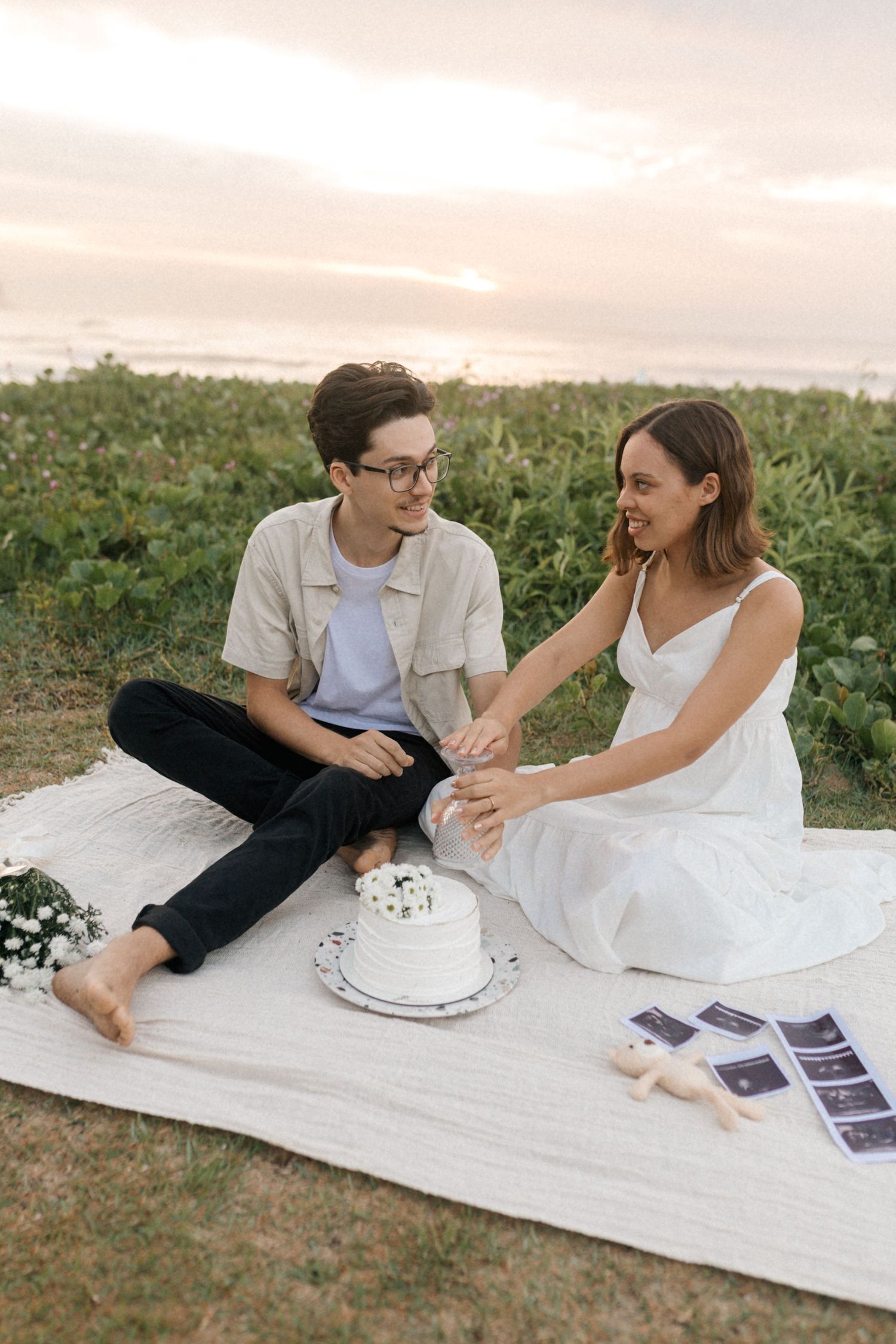 Couple having a picnic on a white blanket while looking at pictures together in a grassy field at sunset.