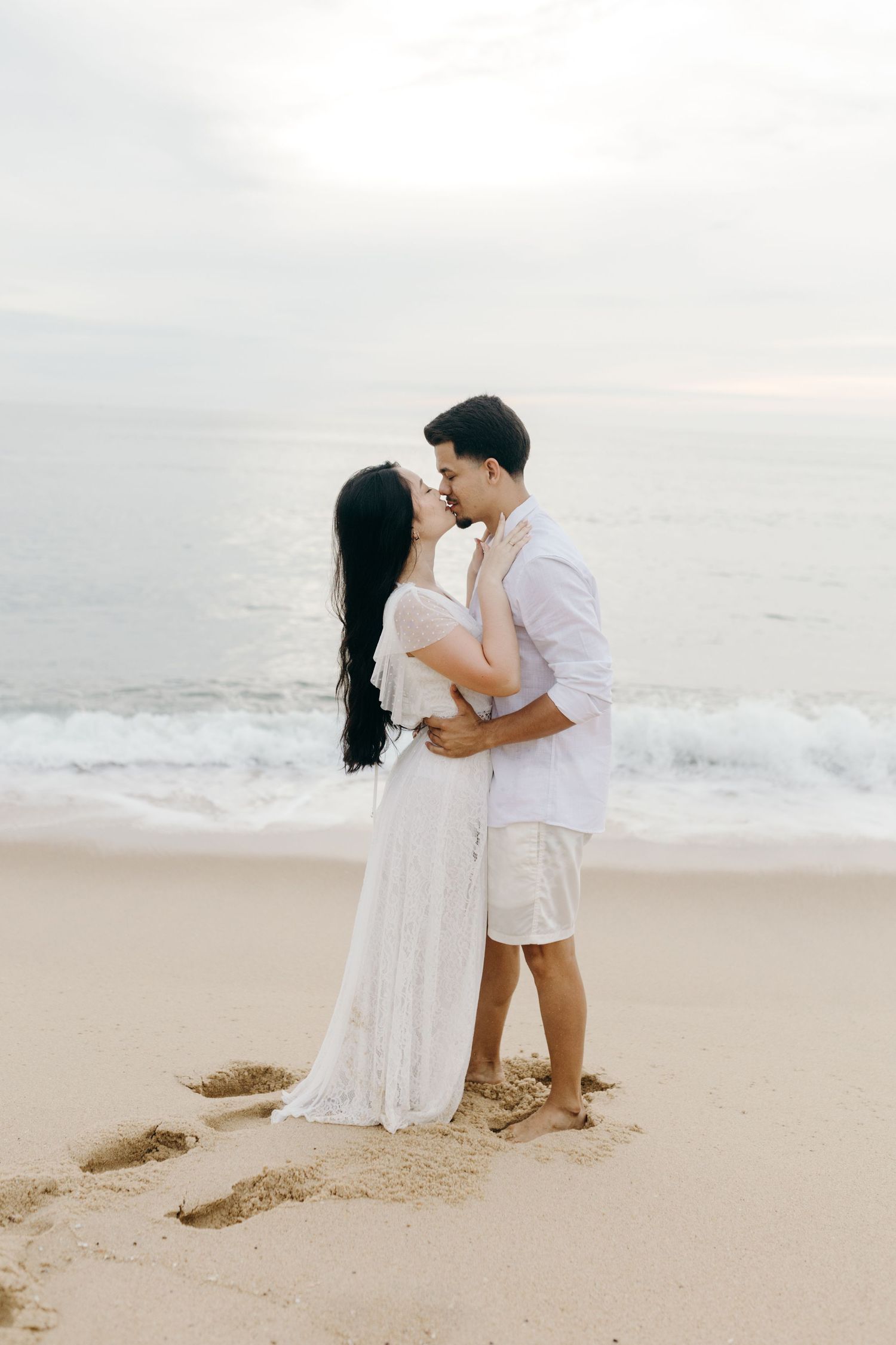 Couple in white attire sharing romantic moments on sandy beach with ocean waves.