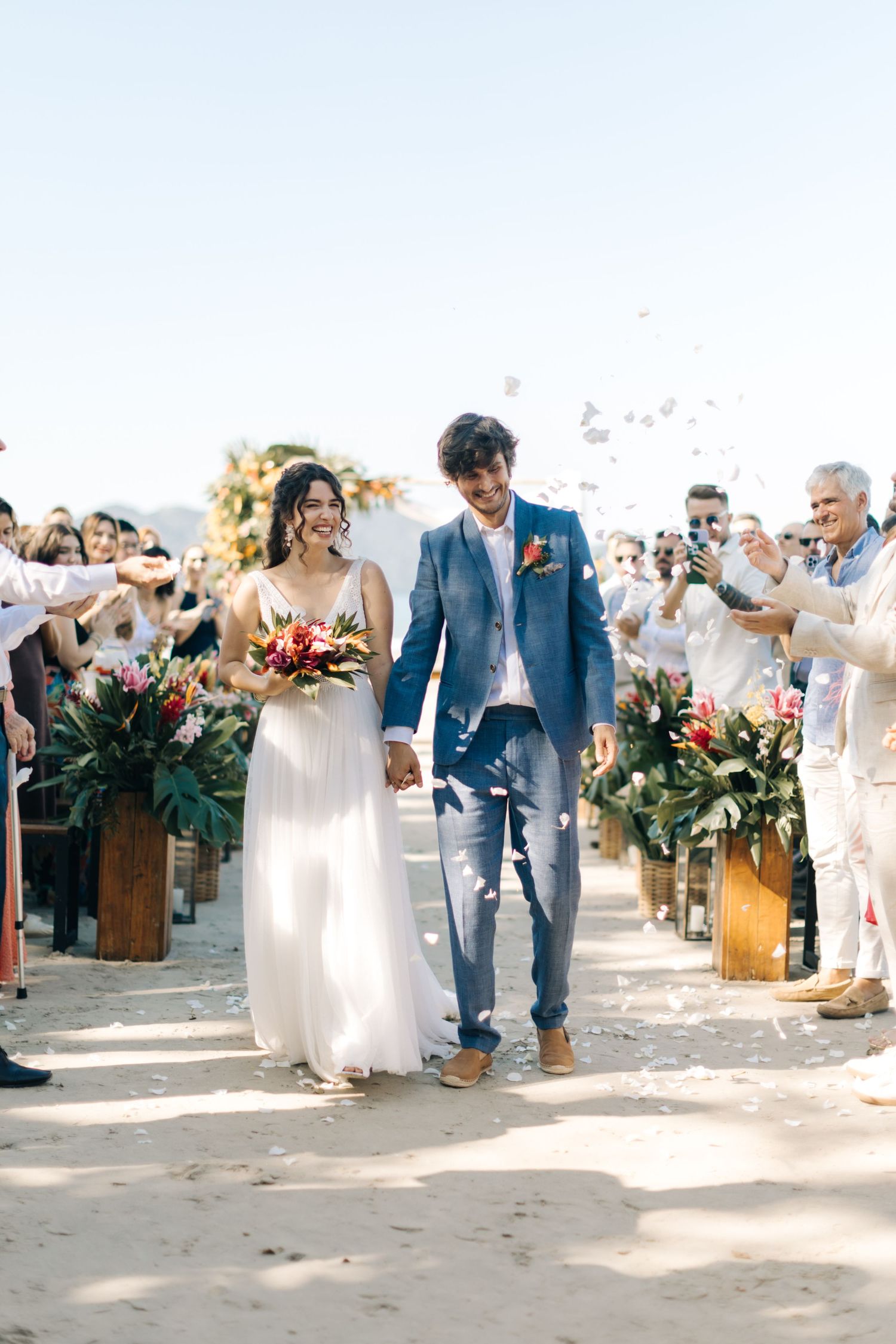 Happy couple walks down sandy aisle lined with wooden planters and cheering guests.